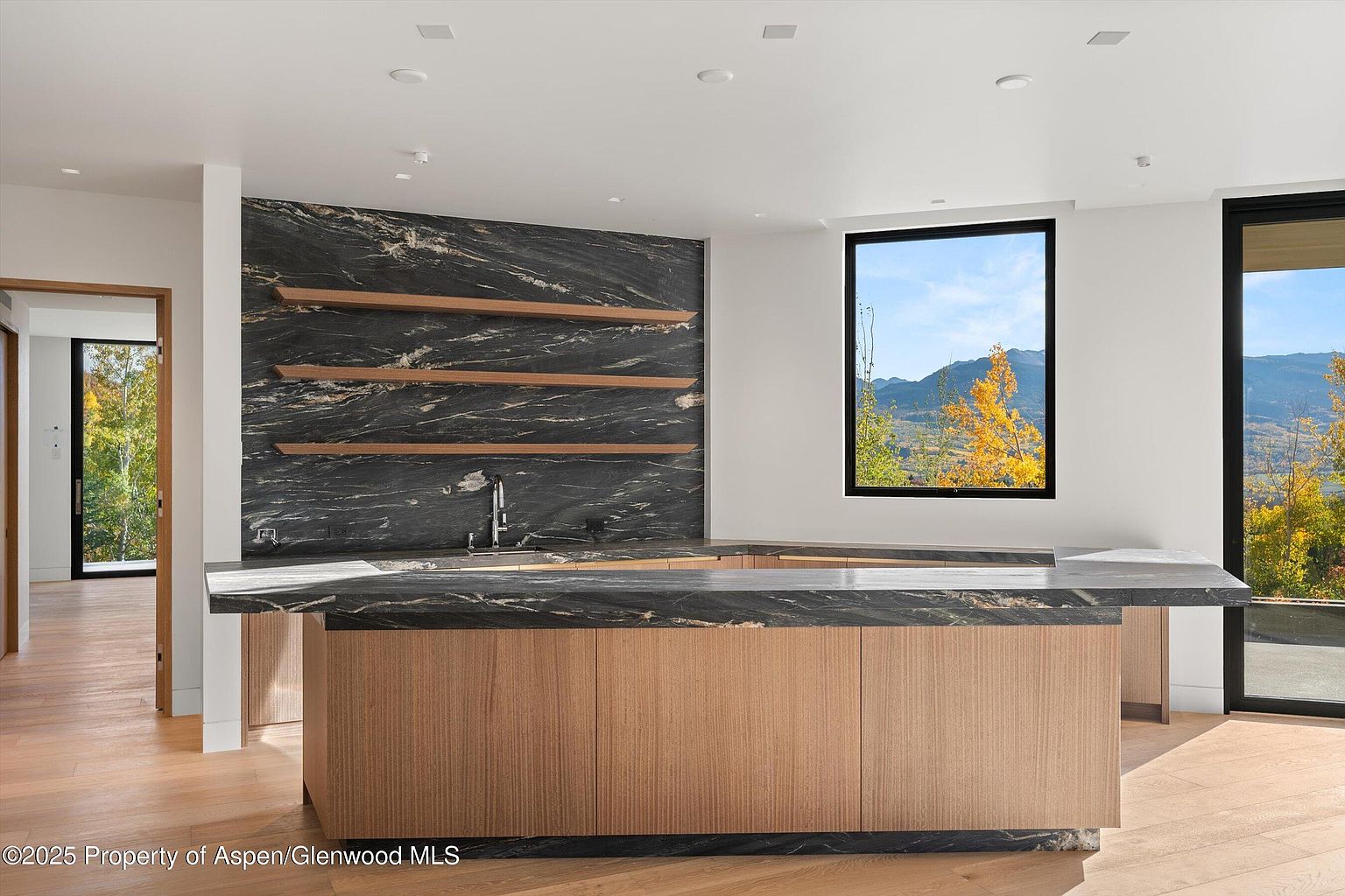 This interior shot showcases a modern kitchen with a striking black marble backsplash and countertop, complemented by light wood cabinetry. Three wooden shelves adorn the marble wall, adding a touch of warmth. A large window provides a scenic view of the outdoors, enhancing the kitchen's bright and airy atmosphere.