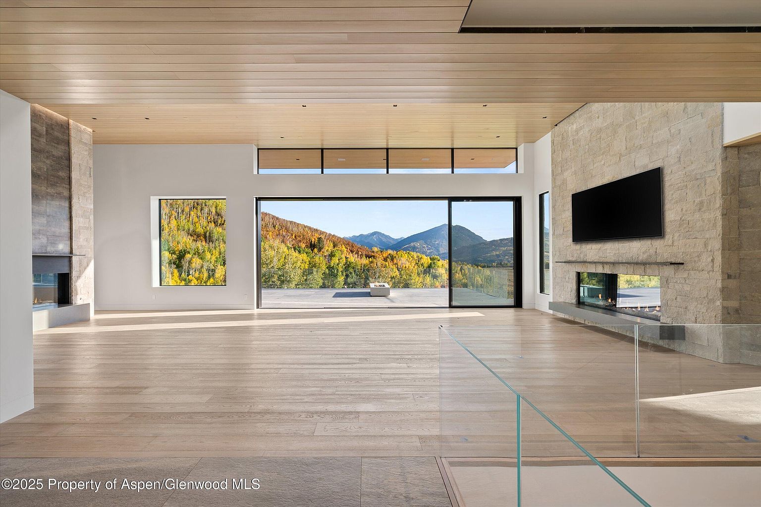 This is a spacious living room featuring hardwood floors, a modern fireplace with a stone surround, and large windows offering a scenic mountain view. The room has a minimalist design with clean lines and a neutral color palette, creating a bright and airy atmosphere. The open floor plan and natural light enhance the sense of space and luxury.