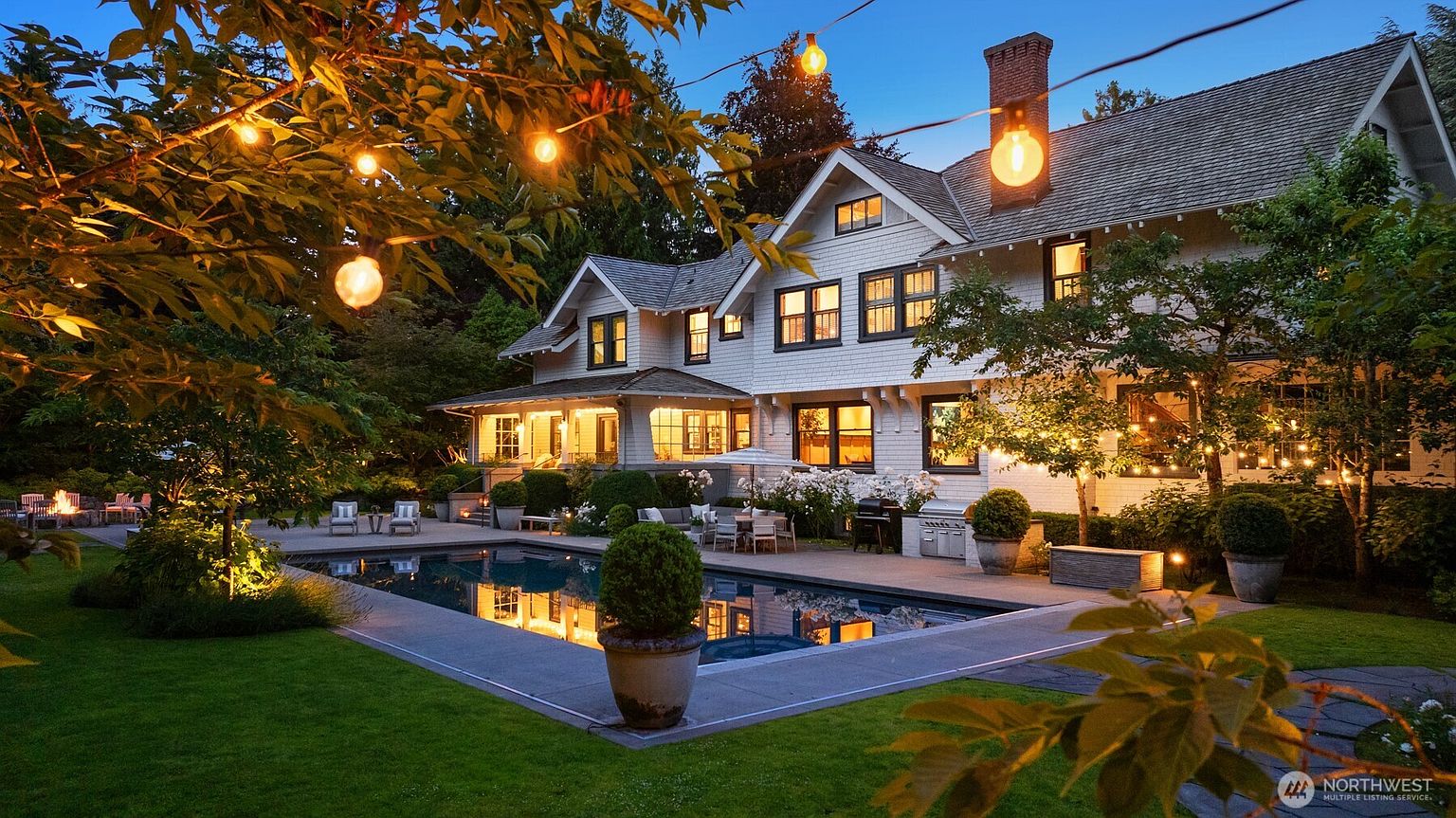 This stunning rear exterior view captures a luxurious multi-story home illuminated at dusk, featuring a pristine rectangular swimming pool as the centerpiece of the backyard. The property boasts a well-manicured lawn, mature landscaping, and a spacious stone patio area perfect for outdoor entertaining, all accented by warm string lighting. The architectural style is elegant and traditional, with large windows reflecting the inviting glow from the interior, creating a sophisticated and serene atmosphere.