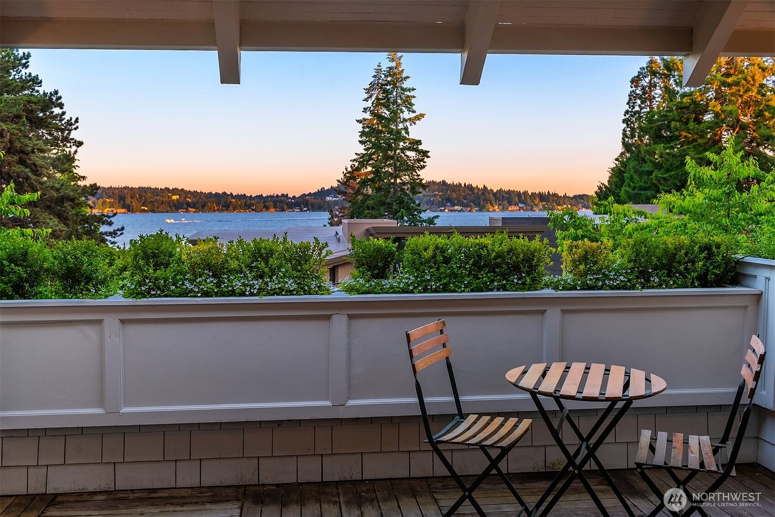 This elevated balcony offers a serene, picturesque view of a calm lake at sunset, framed by lush green trees and a clear, gradient sky. The space is furnished with a simple bistro set, creating an intimate spot for relaxation while overlooking the water and distant shoreline. The wooden decking and white railing provide a clean, classic aesthetic that emphasizes the tranquil outdoor living experience.