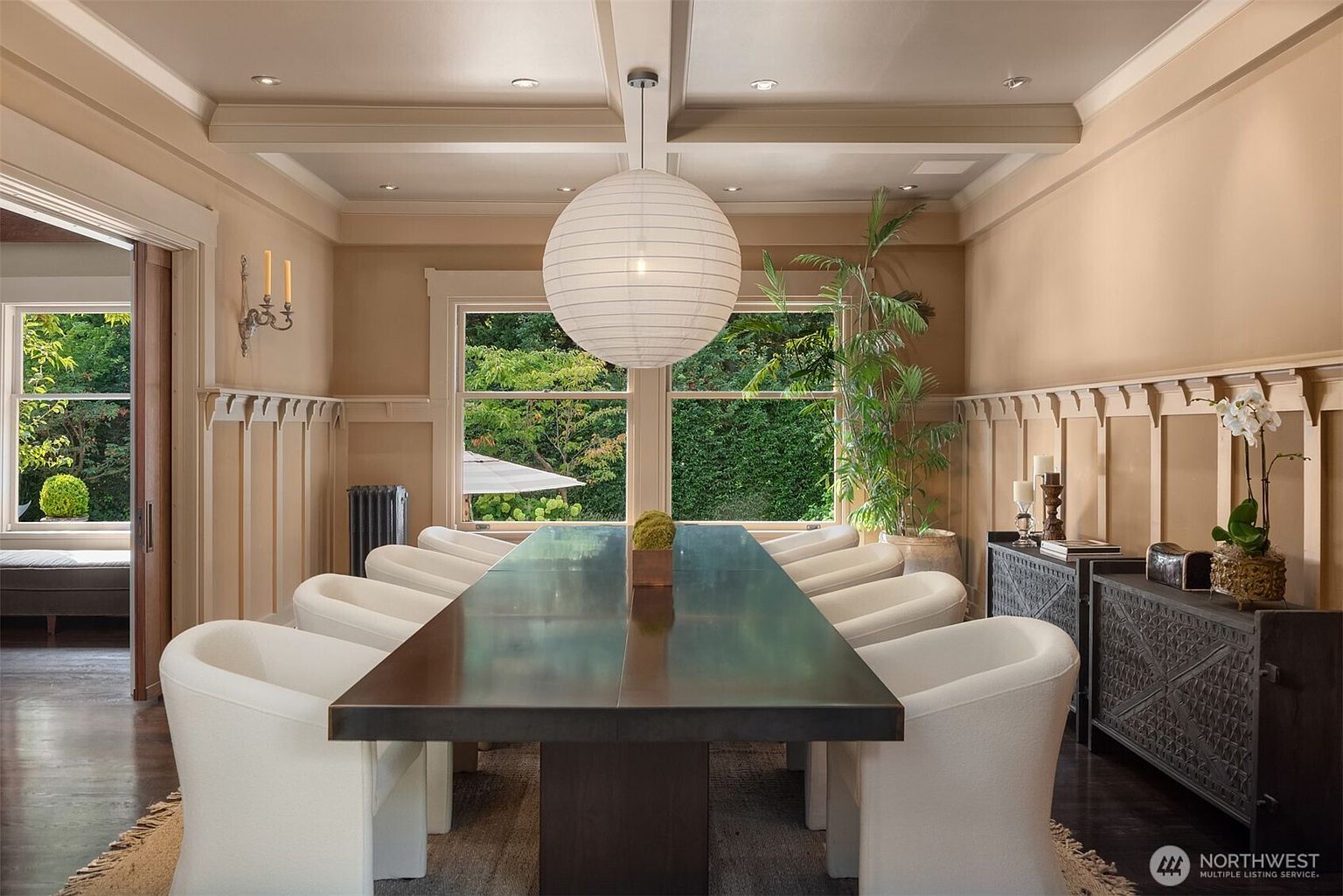 This elegant dining room features a long, dark wood table surrounded by ten modern, cream-colored upholstered chairs, centered under a large, spherical paper pendant light. The room is framed by classic wainscoting and warm-toned walls, with a large window providing a lush, green view of the outdoors. A dark, intricately carved sideboard sits to the right, adding a touch of sophisticated texture to the refined, symmetrical space.