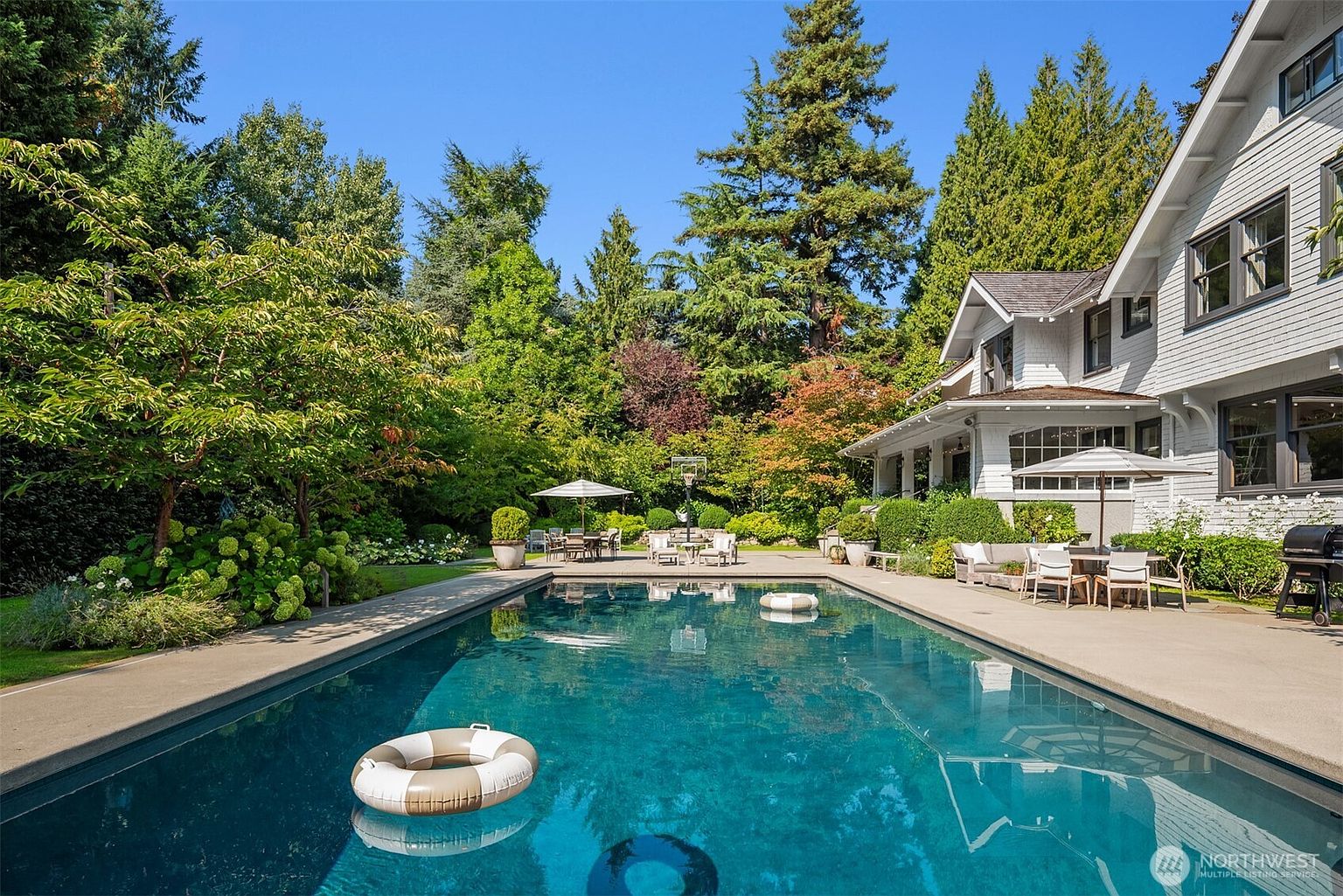 This serene backyard features a large, rectangular swimming pool as the centerpiece, complete with floating rings and a clear, inviting blue surface. The surrounding concrete patio offers ample space for outdoor dining and lounging, framed by lush greenery, mature trees, and a well-manicured garden. The perspective captures the elegant rear facade of the white-sided home, emphasizing a peaceful and luxurious outdoor living environment.