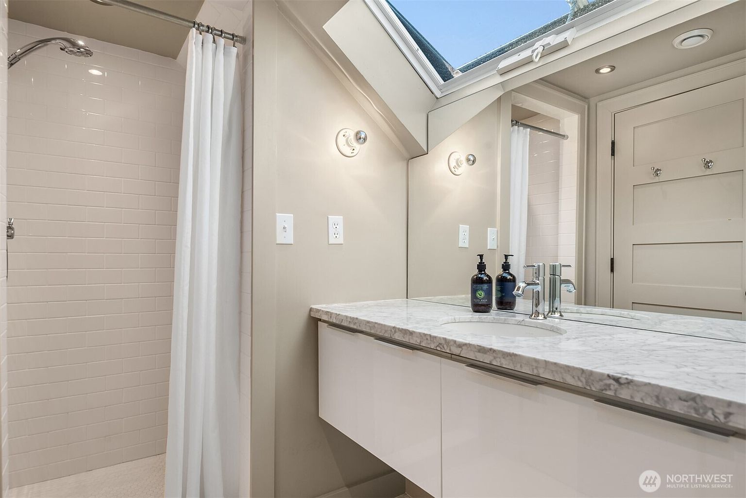 This modern, bright bathroom features a sleek white floating vanity topped with a luxurious marble countertop and a large, wall-to-wall mirror. A walk-in shower with white subway tile is positioned to the left, while a unique skylight above the vanity floods the space with natural light. The overall aesthetic is clean, contemporary, and airy, creating a spa-like atmosphere.
