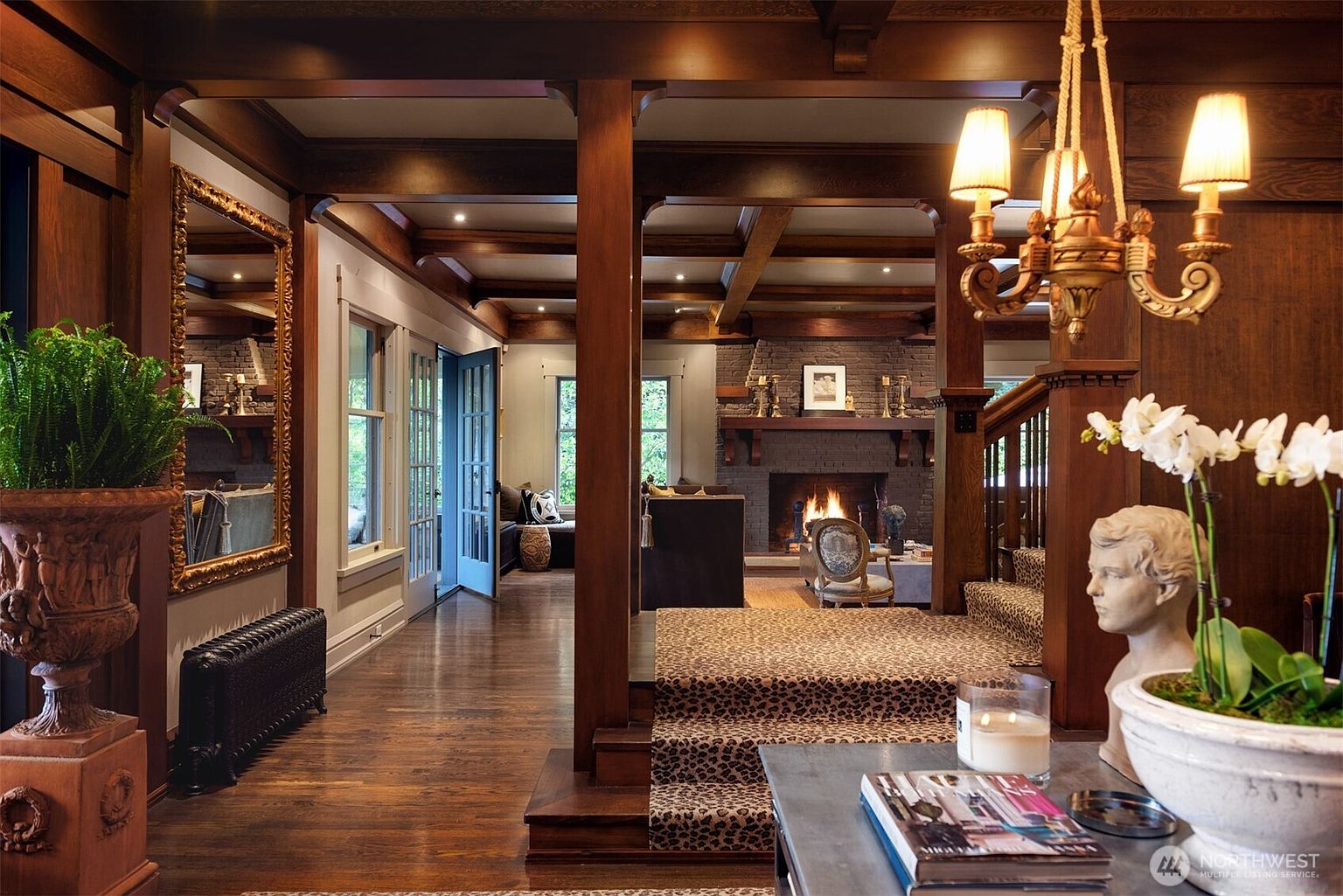 This elegant entryway and hallway transition into a cozy living area, showcasing rich dark wood paneling, exposed ceiling beams, and a warm, inviting fireplace. The space features a sophisticated blend of textures, including a leopard-print carpet on the stairs, a classic chandelier, and a decorative bust, creating a luxurious and timeless atmosphere. The perspective captures the depth of the home, drawing the eye from the foreground details through the open architectural framing toward the glowing hearth.
