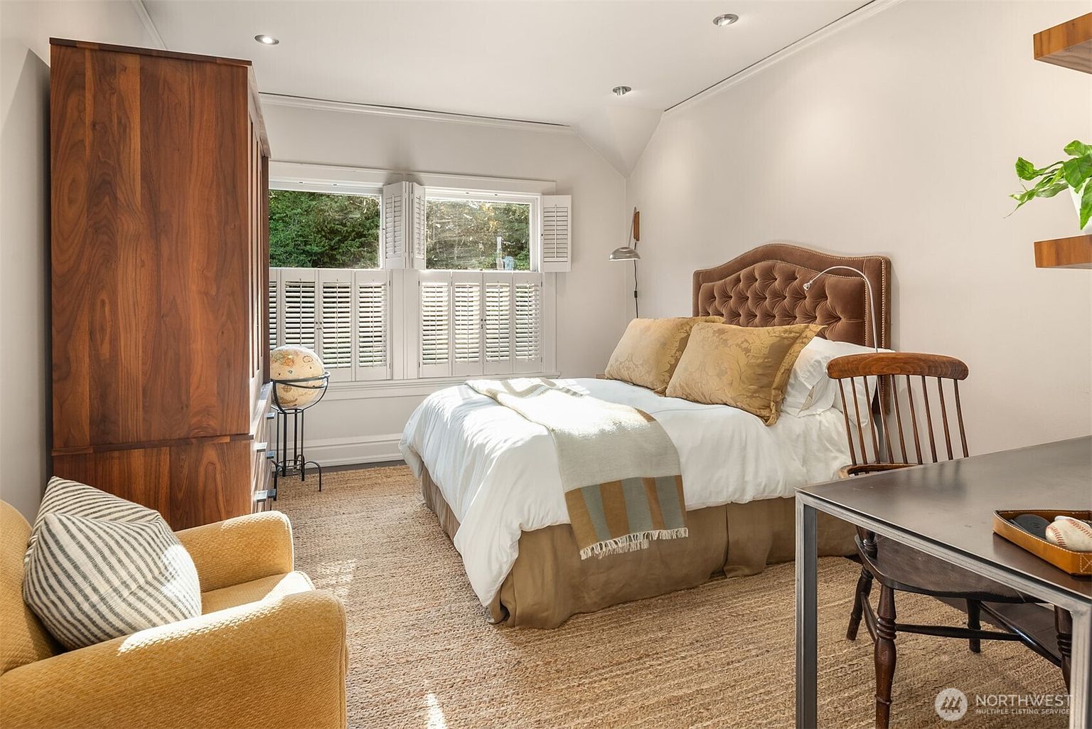 This cozy bedroom features a tufted, brown velvet headboard as the focal point, complemented by a white bedspread and a neutral-toned throw blanket. The room includes a large wooden wardrobe, a mustard-yellow armchair, and a small desk area, all set against a backdrop of light walls and a textured rug. The perspective is from the doorway, capturing the warm, inviting atmosphere and the natural light streaming through the shuttered windows.