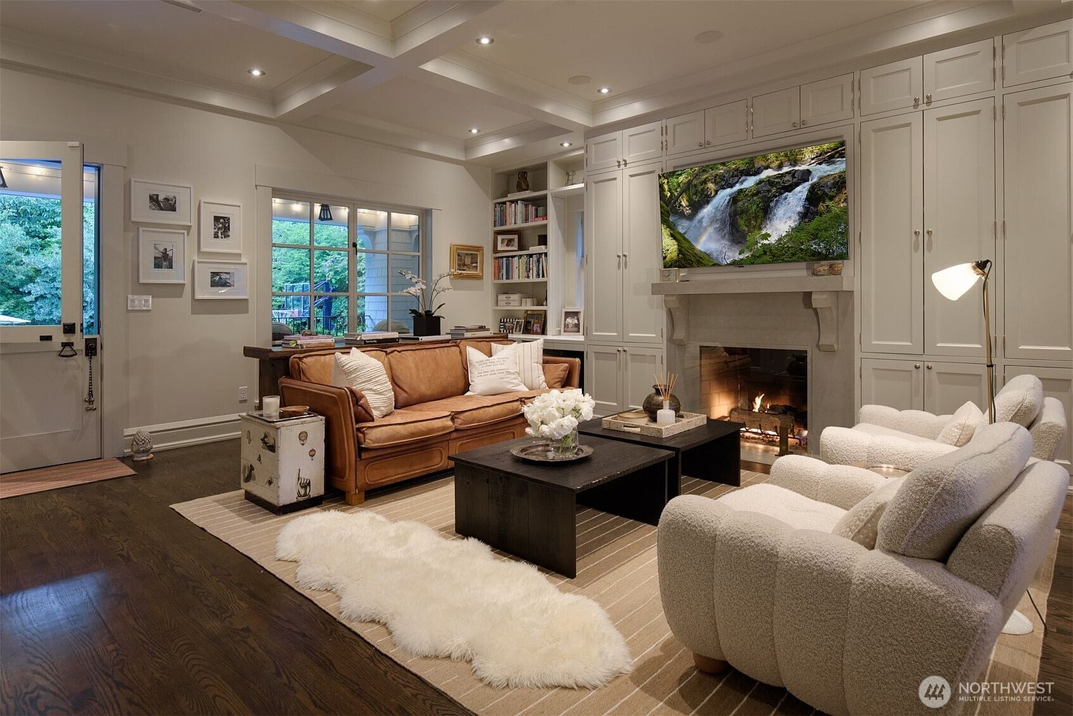 This inviting living room features a warm, sophisticated aesthetic with a rich brown leather sofa, plush cream-colored armchairs, and a cozy faux-fur rug layered over a neutral area rug. A built-in wall unit with integrated shelving and cabinetry surrounds a fireplace topped with a large television, creating a seamless focal point. The room is bathed in natural light from the adjacent window and glass-paneled door, offering a comfortable and elegant space perfect for relaxation.
