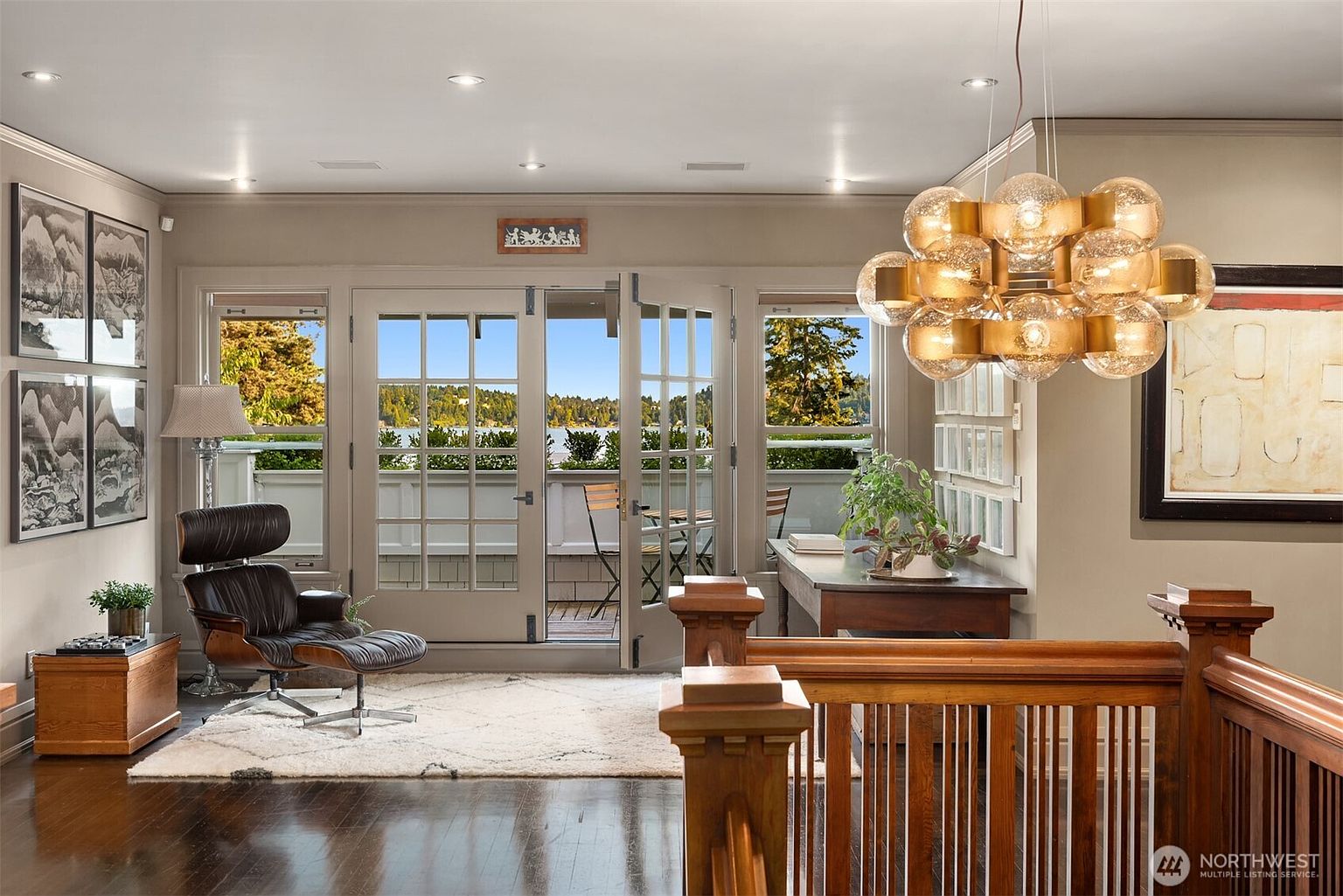 This elegant living space features a classic Eames-style lounge chair set against a backdrop of French doors that open onto a scenic balcony overlooking the water. The room is anchored by a plush white area rug and illuminated by a striking, multi-globe brass chandelier, creating a sophisticated and inviting atmosphere. The warm wood tones of the staircase railing and flooring complement the neutral wall palette, emphasizing a refined, high-end aesthetic.