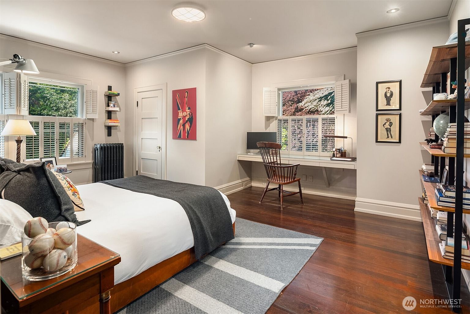 This well-appointed bedroom features rich hardwood flooring, a comfortable bed with a grey throw, and a dedicated built-in desk area under a window with plantation shutters. The room is accented with tasteful decor, including a vintage-style ski poster, framed artwork, and a tall open shelving unit, creating a cozy and functional living space. The perspective is from the doorway, capturing the entire room's layout and inviting atmosphere.