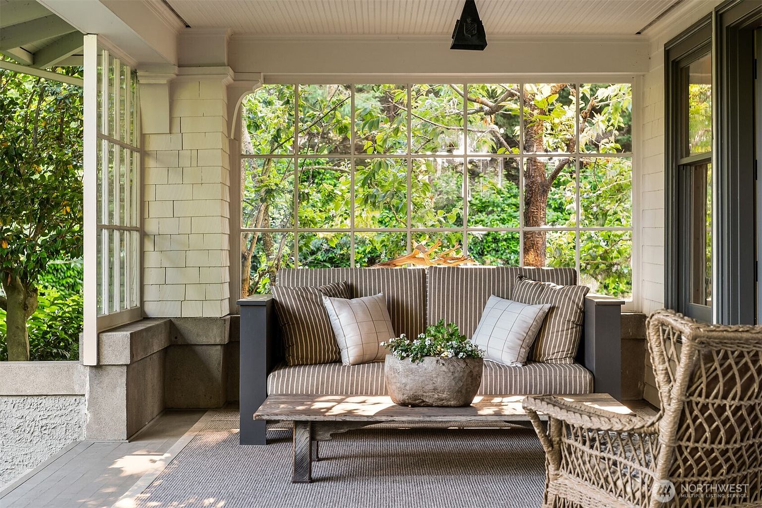 This inviting covered porch features a classic, cozy seating area with a striped sofa and a rustic wooden coffee table topped with a potted plant. The space is enclosed by large, multi-pane windows that offer a serene, leafy view of the surrounding garden, creating a bright and tranquil atmosphere. The combination of natural textures, such as the wicker chair and wooden table, gives the porch a charming, timeless appeal.