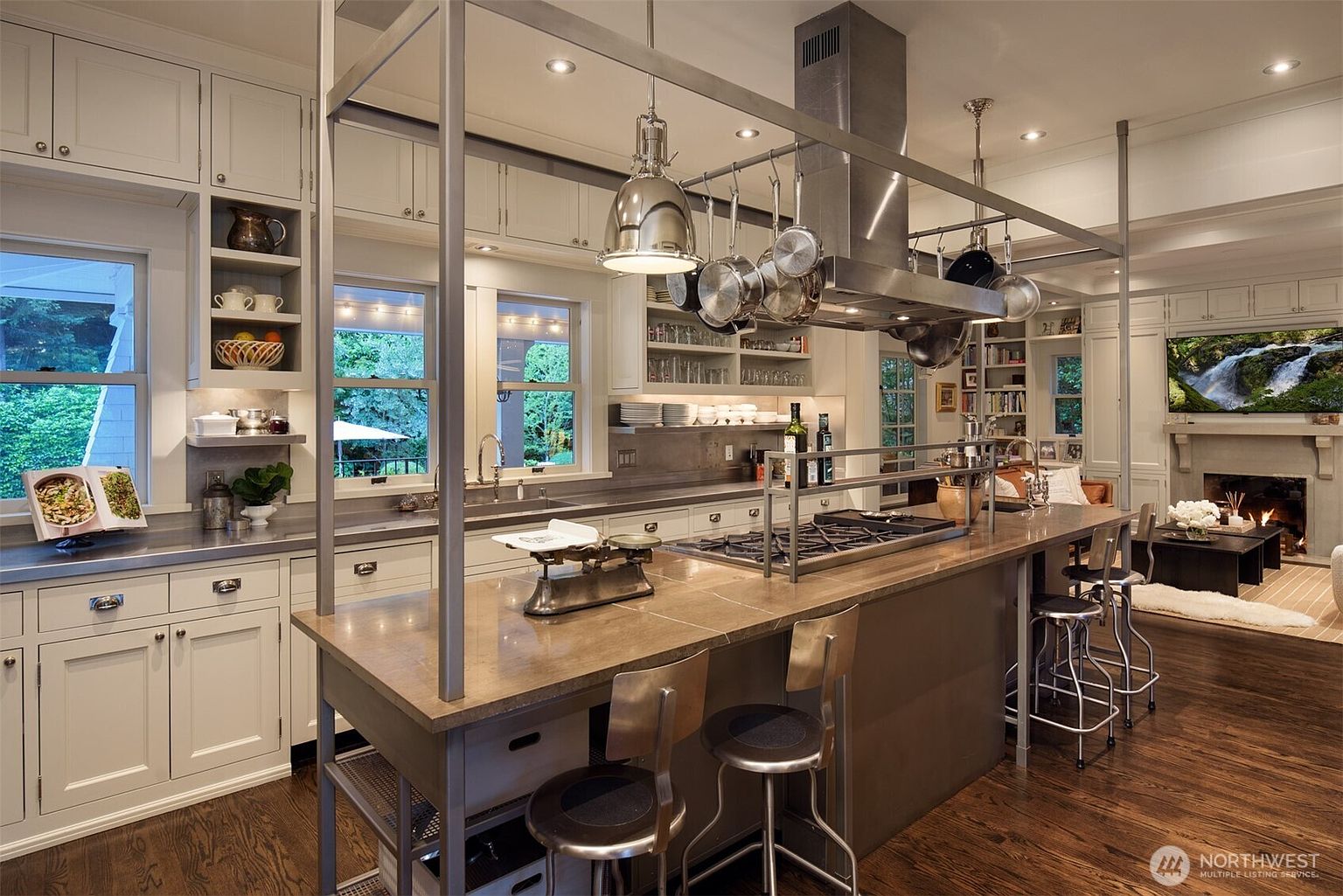 This sophisticated, high-end kitchen features a large central island with a stone countertop, industrial-style metal framing, and bar seating, serving as the focal point of the room. The space is equipped with professional-grade stainless steel appliances, including a prominent range hood, and is accented by white cabinetry and warm wood flooring. The perspective is a wide, eye-level shot that captures the seamless flow between the culinary workspace and the adjacent living area, creating an inviting and open-concept atmosphere.