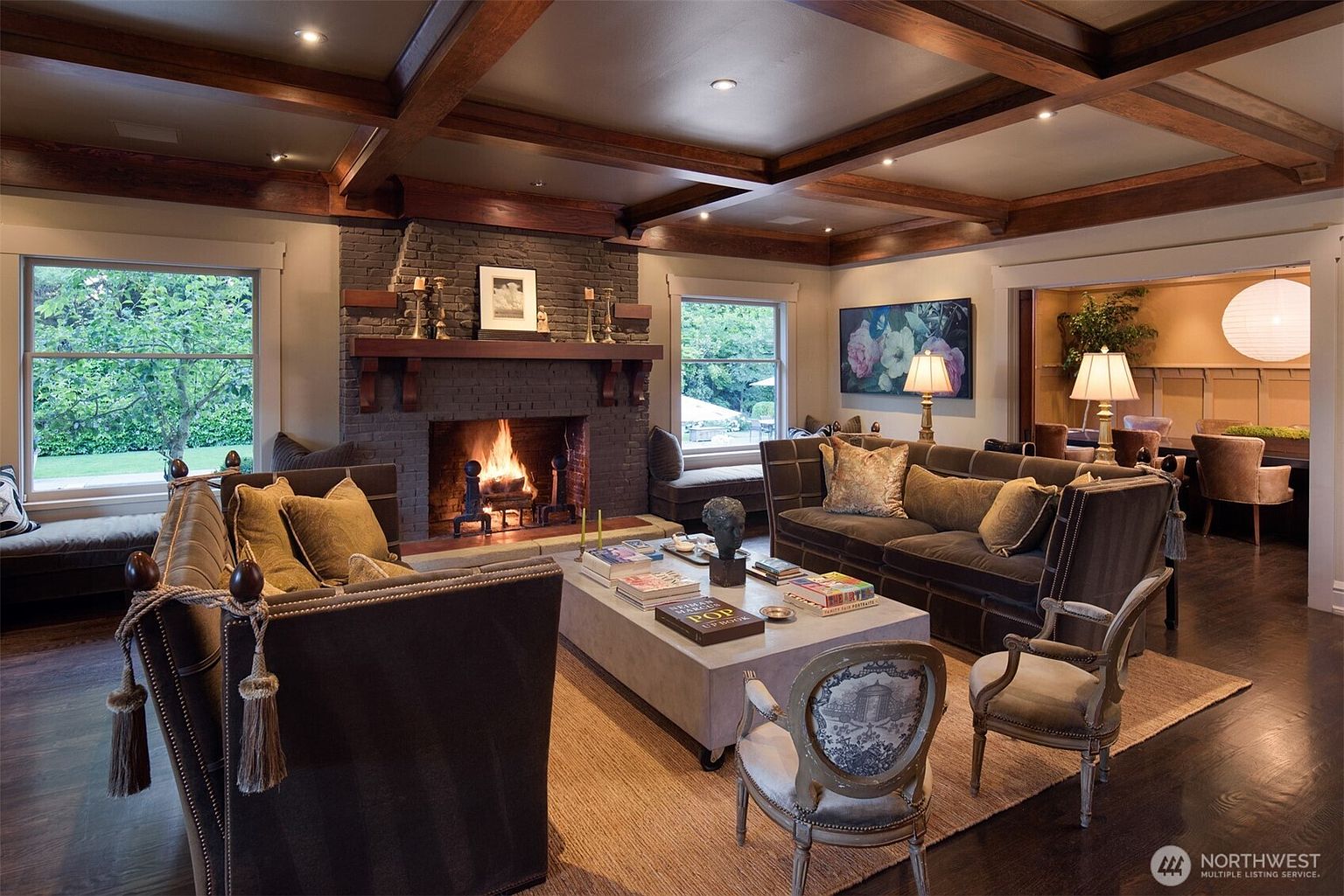 This elegant living room features a dark brick fireplace as its focal point, complemented by rich wood-beamed ceilings and dark hardwood floors. The space is furnished with plush, dark-toned sofas and vintage-style accent chairs arranged around a large, modern concrete coffee table. The warm glow of the fire and soft ambient lighting create a sophisticated and inviting atmosphere, with a glimpse into an adjacent dining area.