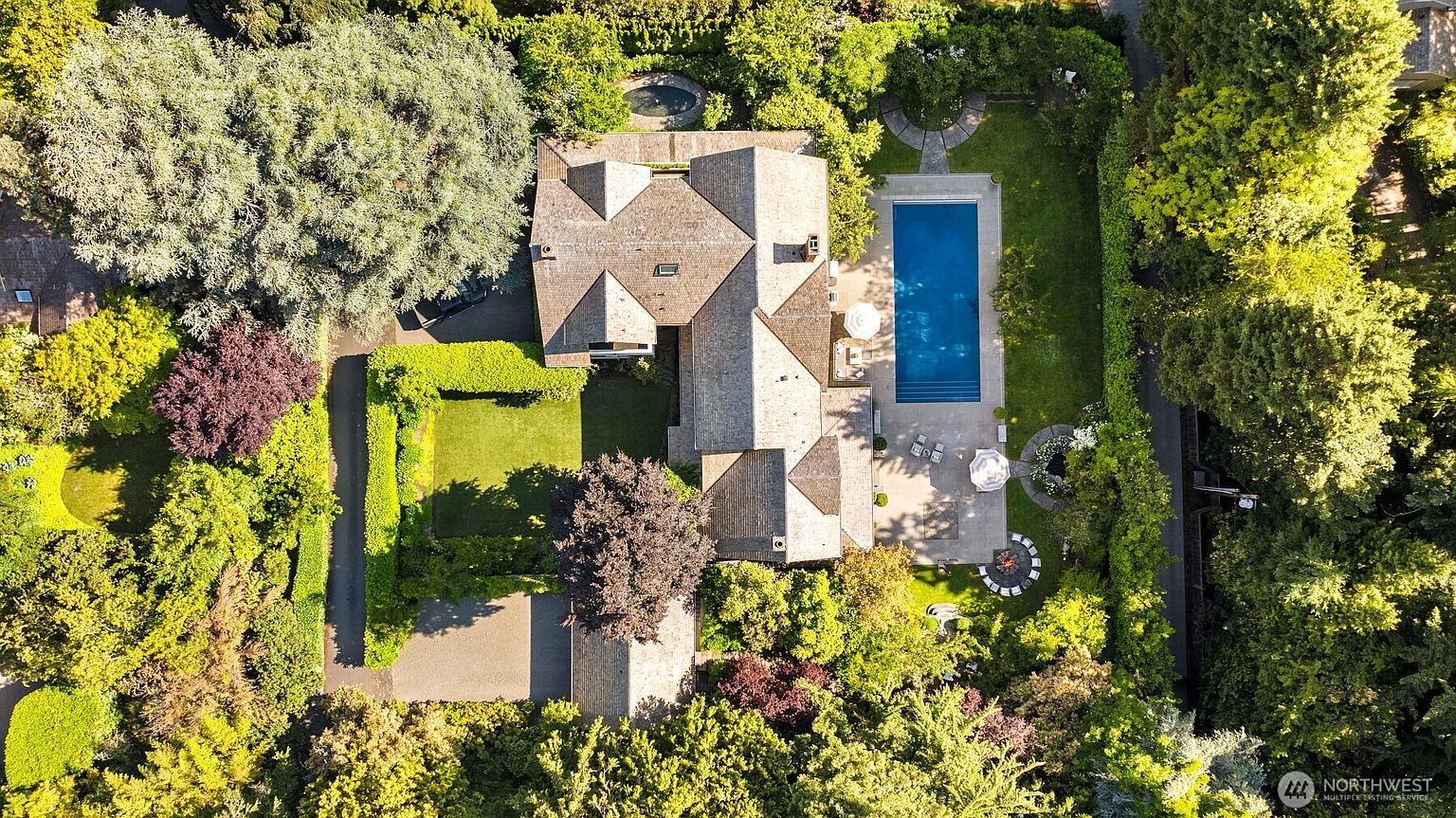This high-angle aerial view captures a sprawling residential property nestled within a lush, mature landscape. The home features a complex, multi-gabled roofline and is complemented by a rectangular swimming pool, a stone patio with a fire pit, and meticulously manicured green lawns. The dense surrounding trees provide significant privacy, creating a serene and secluded estate atmosphere.