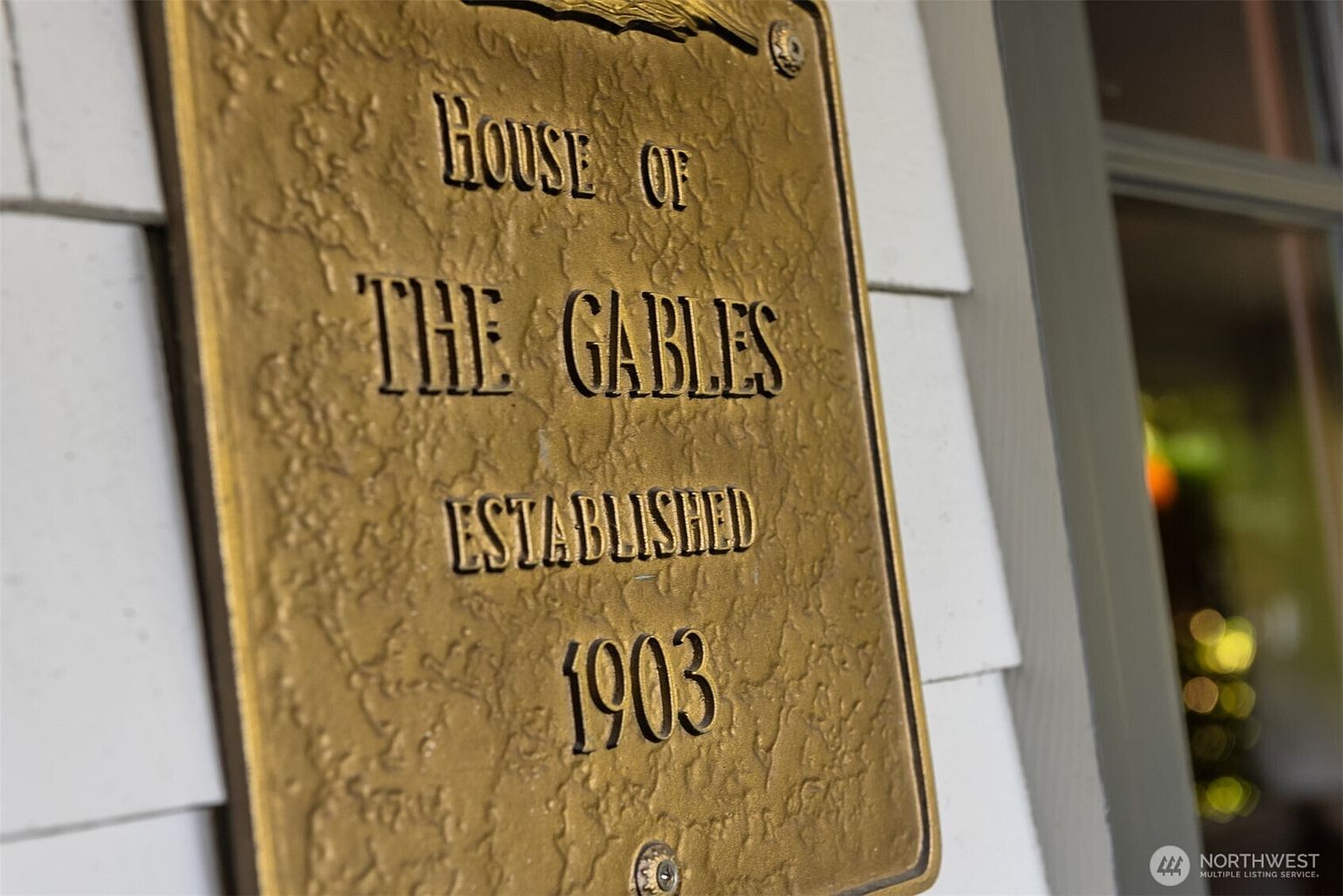 A close-up, angled shot of a textured, gold-toned metal plaque mounted on white shingle siding. The plaque reads 'HOUSE OF THE GABLES' and 'ESTABLISHED 1903' in raised, dark lettering, evoking a sense of historic charm and heritage. The perspective highlights the tactile quality of the sign and its integration into the home's exterior facade.