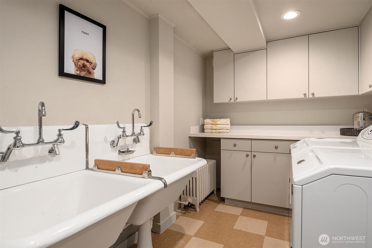 This functional laundry room features a vintage-style double basin utility sink with chrome fixtures and wooden washboards, paired with modern white cabinetry and a washing machine. The space is finished with neutral-toned walls and a checkered floor, creating a clean and practical utility area. A framed portrait of a dog adds a personalized touch to the room, which is illuminated by recessed ceiling lighting.