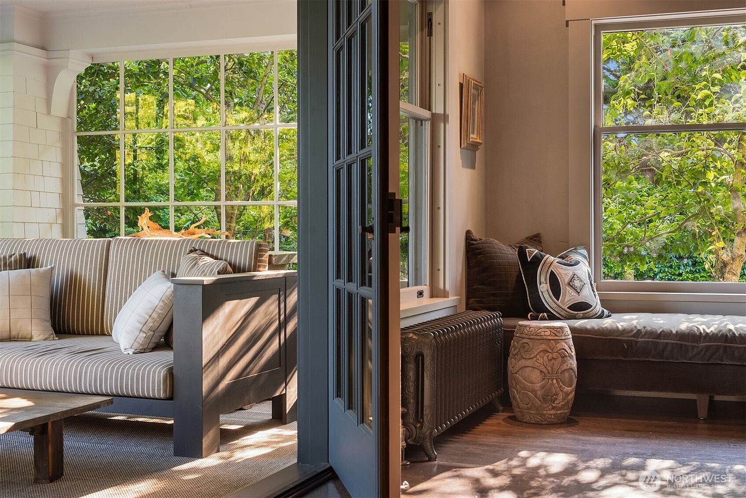 This split-view composition captures a cozy sunroom transition, featuring a striped sofa on the left and a comfortable window seat on the right. The space is bathed in natural light from large, multi-pane windows that frame lush green foliage, creating a serene and inviting atmosphere. The contrast between the dark, classic radiator and the soft, textured upholstery highlights a blend of traditional charm and relaxed comfort.