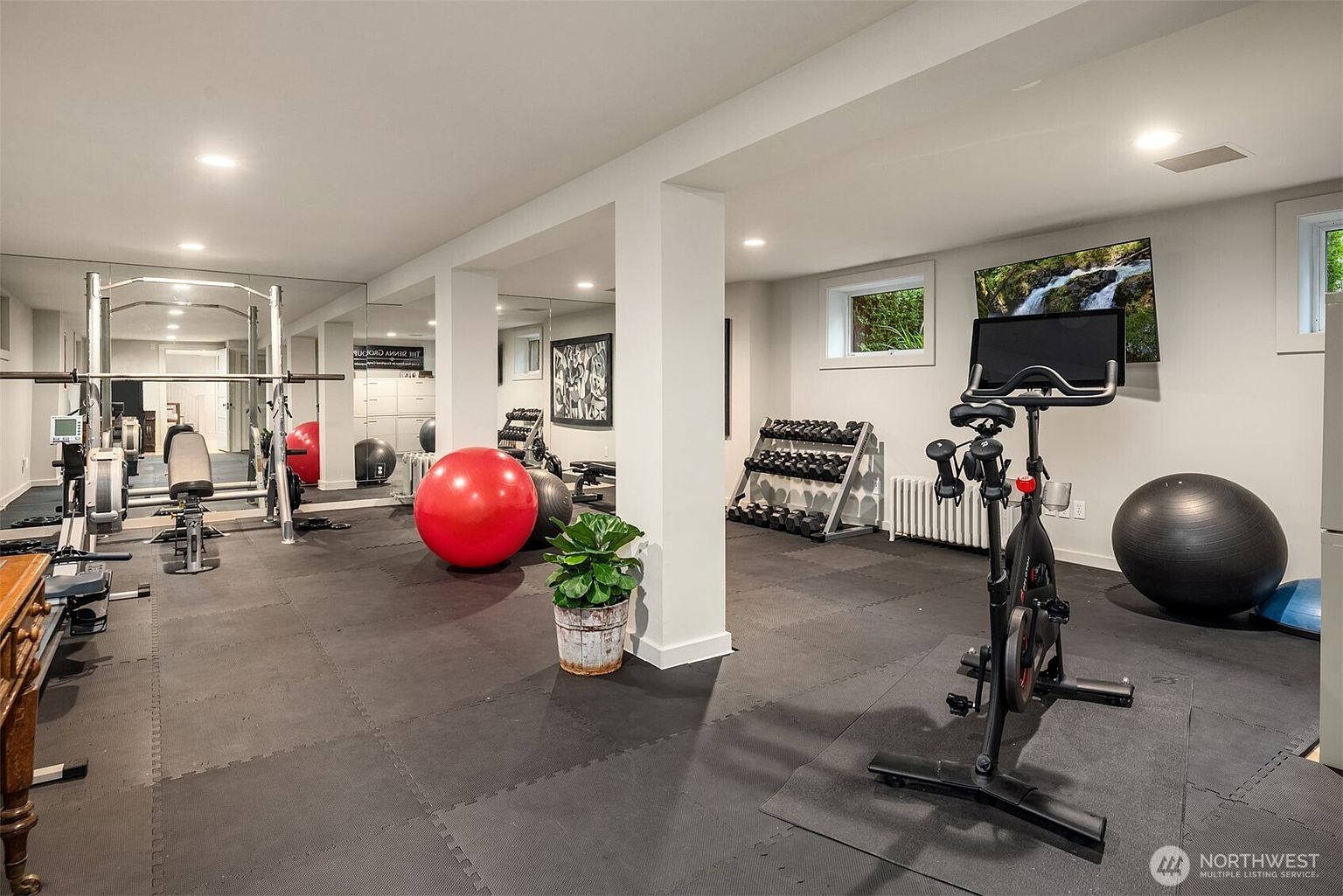 This spacious, well-lit home gym features professional-grade equipment including a stationary bike, a weight rack with dumbbells, and a squat rack. The room is finished with durable black rubber flooring, mirrored walls to enhance the sense of space, and a wall-mounted television for entertainment during workouts. The clean, neutral color palette and functional layout create an inviting and motivating environment for fitness enthusiasts.