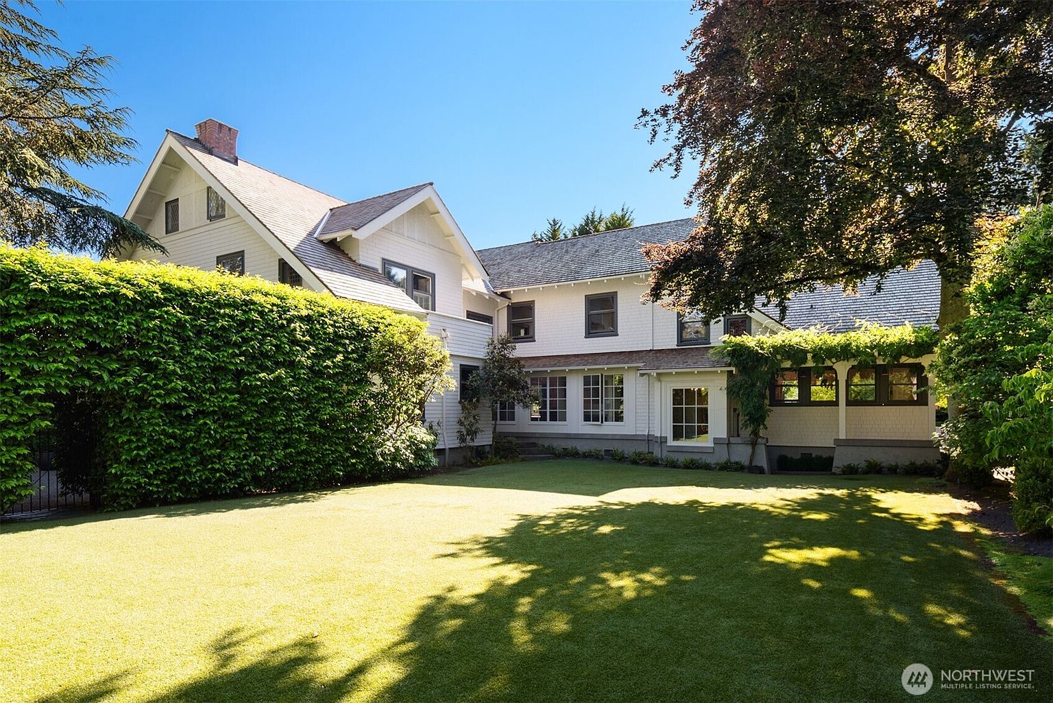 This image captures the rear exterior of a classic, multi-story white shingle-style home, showcasing its architectural charm and expansive backyard. The property features a well-manicured lawn, a lush privacy hedge, and a pergola-covered patio area that seamlessly blends indoor and outdoor living. The perspective is a wide, eye-level shot that emphasizes the home's scale and the serene, park-like setting of the garden.