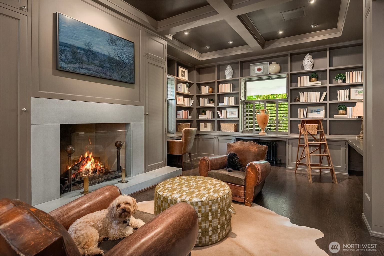 This sophisticated home office or library features rich, dark-toned built-in shelving, a coffered ceiling, and a sleek stone fireplace that serves as a focal point. The room is furnished with two comfortable leather armchairs, a patterned ottoman, and a wooden library ladder, creating a cozy and scholarly atmosphere. The perspective is a wide-angle shot from the doorway, capturing the warmth of the fire and the inviting, well-appointed layout.