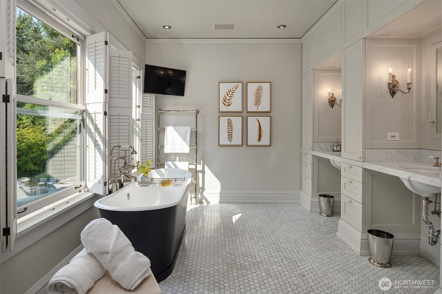 This elegant primary bathroom features a classic clawfoot tub as the centerpiece, positioned next to a large window with plantation shutters that flood the space with natural light. The room is finished with white hexagonal floor tiles, dual vanity stations with marble countertops, and sophisticated wall sconces. A wall-mounted television and framed botanical art add a touch of modern luxury to the serene, neutral-toned space.