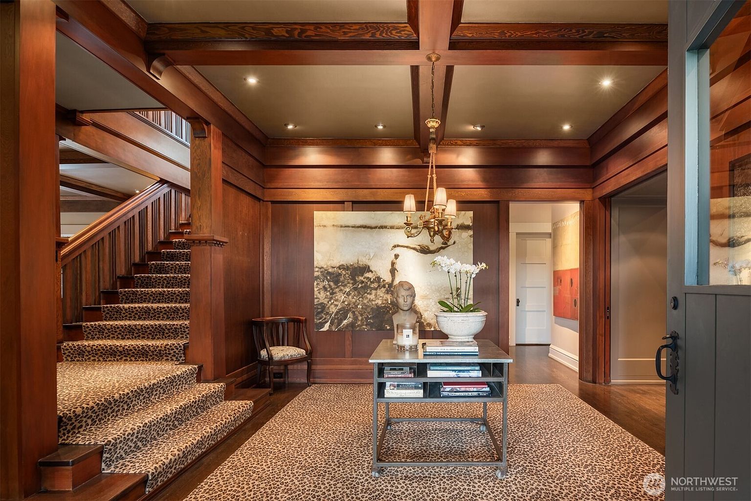 This grand foyer features rich, dark wood paneling and exposed ceiling beams, creating a sophisticated and warm atmosphere. A striking staircase with a leopard-print runner leads to the upper level, while a central metal console table and a classic chandelier serve as elegant focal points. The space is anchored by a matching leopard-print area rug, blending traditional architectural craftsmanship with bold, modern design elements.
