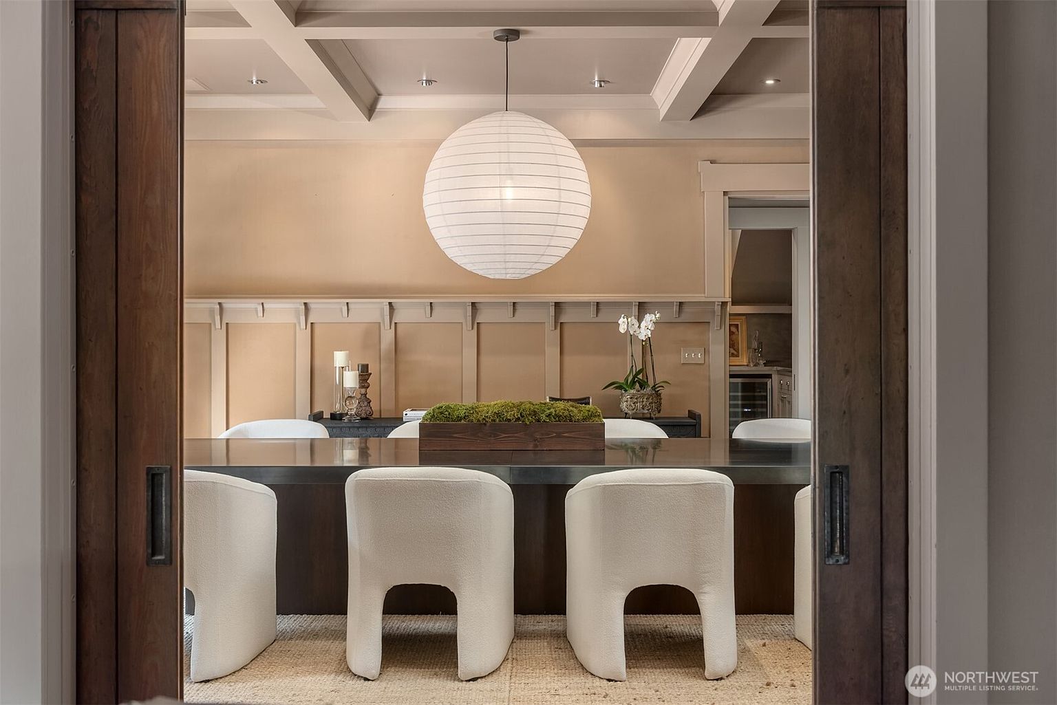 This elegant dining room is framed by dark wood sliding doors, creating a sophisticated and inviting perspective. The space features a long, dark wood table topped with a moss-filled centerpiece, surrounded by modern, cream-colored upholstered chairs, all illuminated by a large, spherical paper pendant light. The room is accented by classic wainscoting and a coffered ceiling, exuding a refined and polished aesthetic.