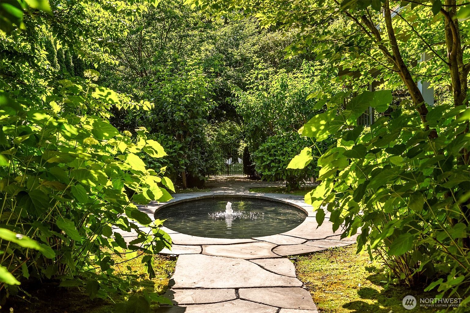 This serene garden scene features a circular stone-rimmed fountain as the focal point, set amidst lush, mature greenery and trees. A flagstone path leads directly to the water feature, creating a tranquil and inviting atmosphere. The perspective is eye-level, capturing the dappled sunlight filtering through the dense foliage, which enhances the private and peaceful feel of the landscape.