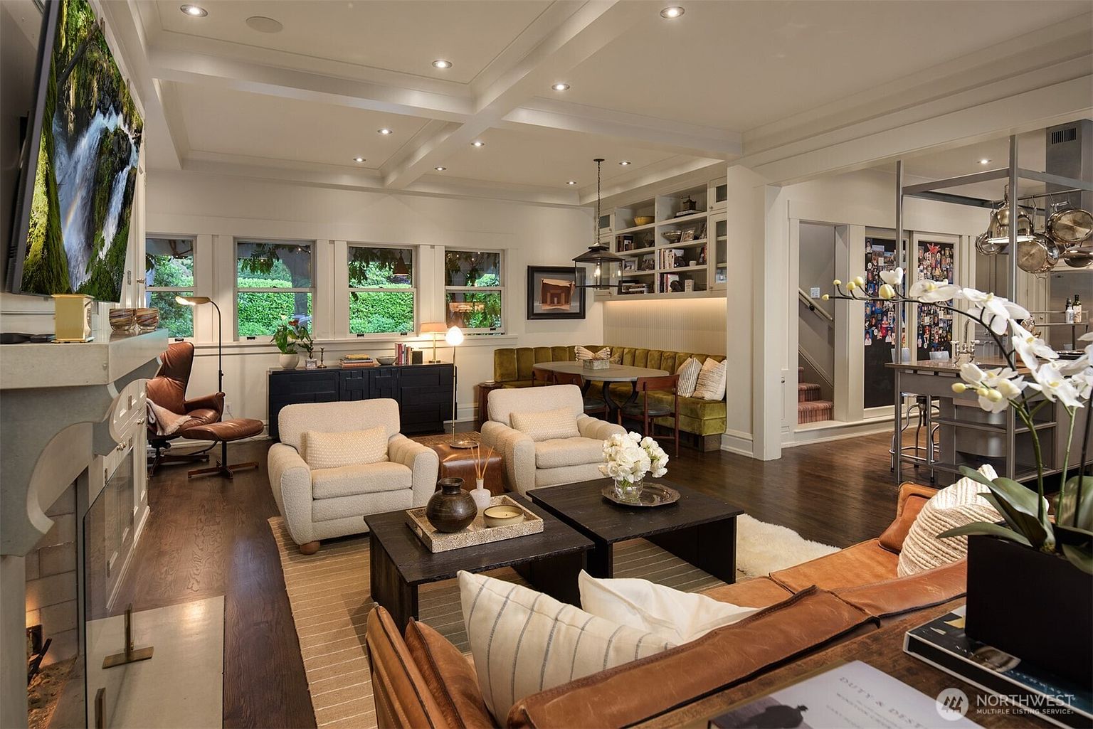 This inviting open-concept living space features a warm, sophisticated design with dark hardwood floors, a cozy leather sofa, and two plush cream armchairs centered around dual coffee tables. A built-in dining nook with a green velvet banquette and custom shelving adds character, while the coffered ceiling and large windows create an airy, high-end atmosphere. The perspective is from the foreground, looking past the sofa toward the fireplace and dining area, emphasizing the seamless flow of the home's layout.