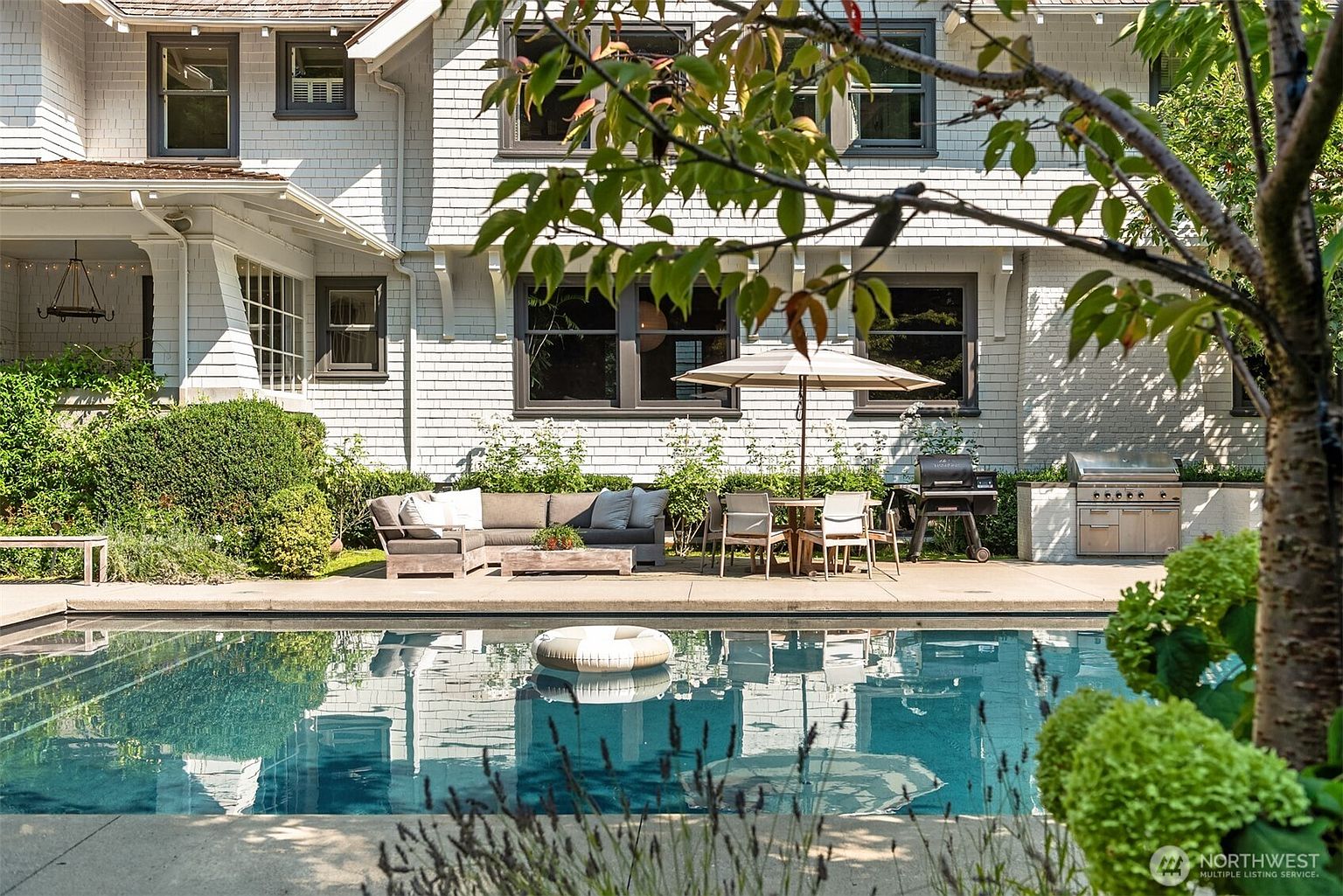 This inviting backyard scene features a pristine rectangular swimming pool in the foreground, complete with a floating ring. The patio area is well-appointed with a comfortable sectional sofa, a dining set shaded by an umbrella, and a built-in outdoor kitchen with a grill. The white-shingled house provides a classic, elegant backdrop, creating a serene and luxurious atmosphere perfect for outdoor entertaining.