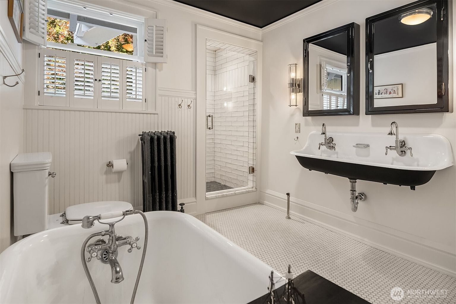 This elegant primary bathroom features a classic clawfoot tub, a double-basin wall-mounted sink, and a separate glass-enclosed shower with subway tile. The room is styled with a sophisticated black-and-white color palette, including a dark ceiling, black-framed mirrors, and a vintage-inspired radiator. The perspective is a wide-angle shot that captures the entire layout, emphasizing the blend of historic charm and modern luxury.