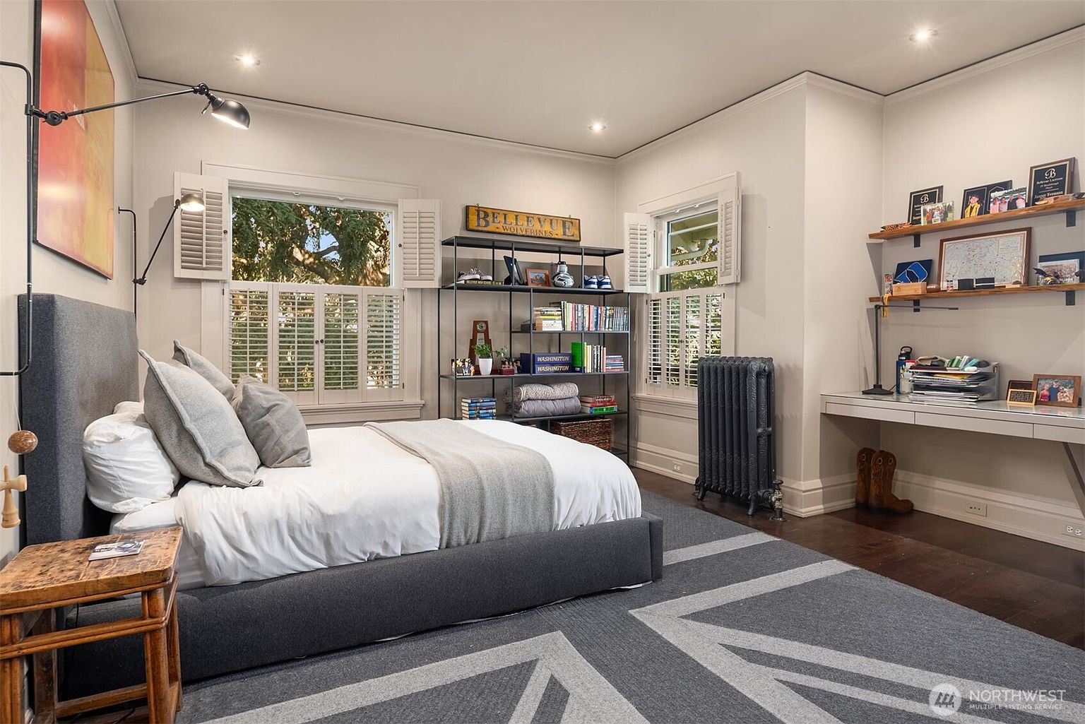 This well-appointed bedroom features a modern gray upholstered bed frame centered against a wall with a large piece of abstract art and a sleek wall-mounted lamp. The room includes a dedicated study area with a floating desk and open shelving, complemented by a tall metal bookshelf and a vintage-style radiator. Large windows with white plantation shutters allow natural light to fill the space, creating a cozy and functional atmosphere.