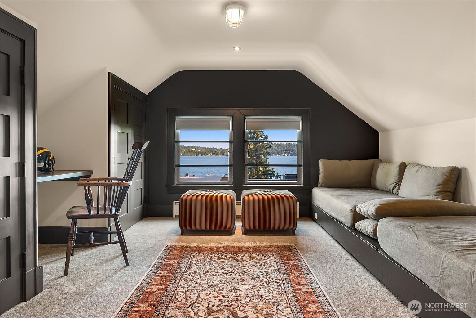 This cozy attic space features a sloped ceiling, dark accent walls, and a large window offering a serene view of the lake. The room is furnished with a comfortable daybed, two striped ottomans, and a classic wooden chair at a built-in desk, all set atop a patterned area rug. The overall atmosphere is quiet and contemplative, making it an ideal retreat or home office.