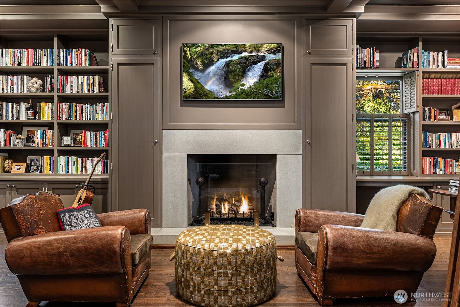 This sophisticated study features floor-to-ceiling built-in bookshelves flanking a central fireplace with a stone surround and a mounted television. Two distressed leather armchairs and a patterned ottoman create a cozy seating area, while the warm wood flooring and dark cabinetry evoke a classic, scholarly atmosphere. The room is framed symmetrically, offering a balanced and inviting perspective perfect for reading or quiet work.