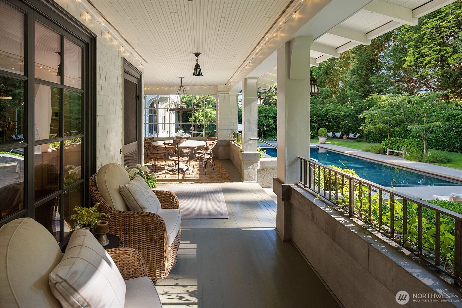 This inviting covered porch features comfortable wicker seating and a dining area, creating a perfect space for outdoor relaxation and entertaining. The space overlooks a serene swimming pool and lush, manicured garden, blending classic architectural charm with a tranquil resort-like atmosphere. The perspective captures the seamless transition between the shaded living area and the sun-drenched backyard.
