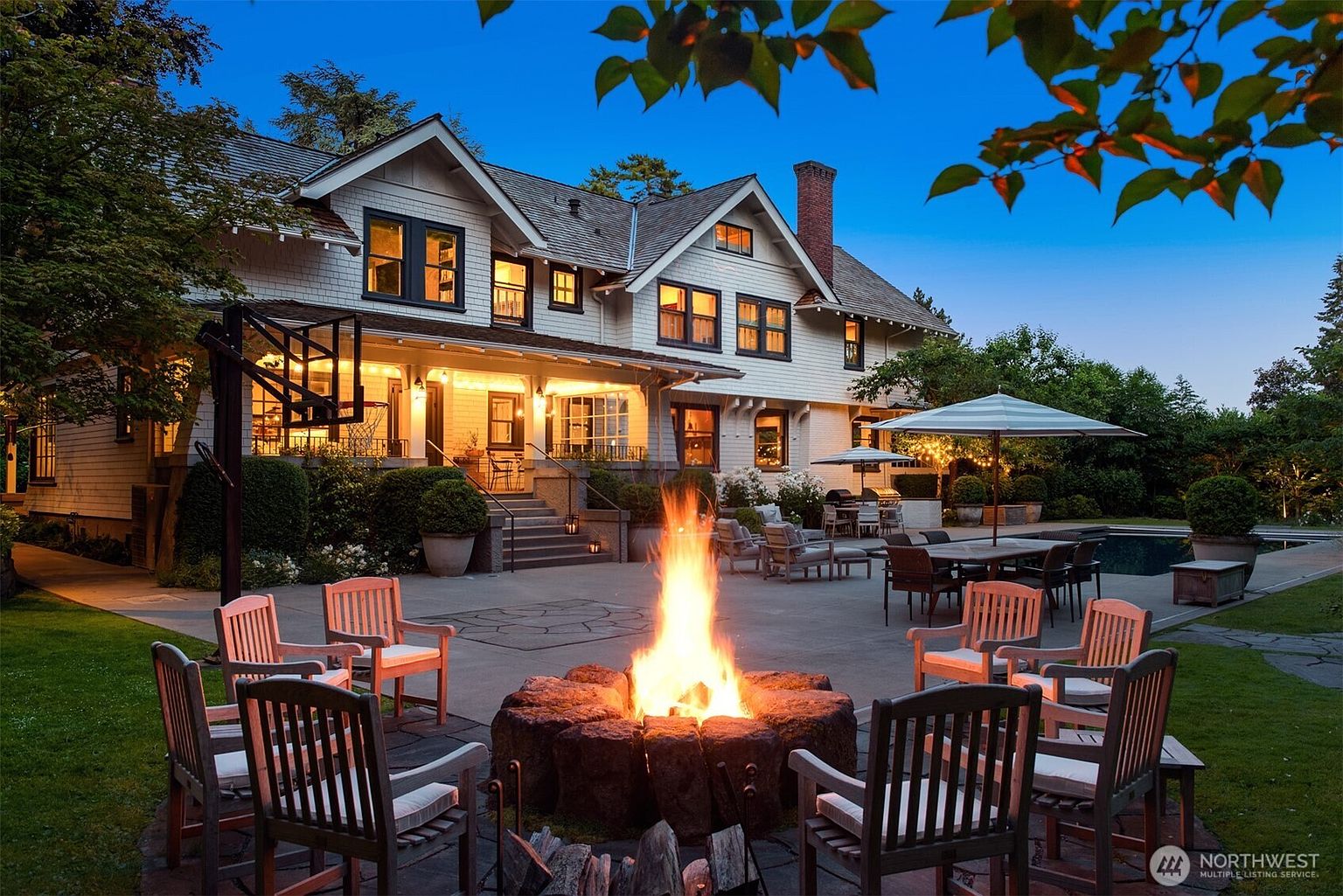 This stunning rear exterior view captures a grand, traditional-style home at dusk, featuring a spacious stone patio centered around a large, rustic stone fire pit. The scene is beautifully illuminated by warm ambient lighting from the house and string lights, highlighting a dining area, a swimming pool, and a basketball hoop. The perspective is low and cinematic, emphasizing the inviting, luxurious atmosphere of the outdoor living space.