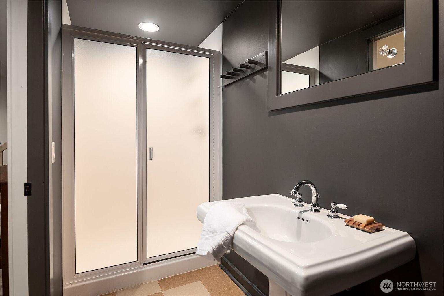 This modern bathroom features a sleek, dark-painted wall that contrasts with a white pedestal sink and a frosted glass shower enclosure. A wall-mounted coat rack and a framed mirror add functional, minimalist style to the compact space. The overall aesthetic is contemporary and moody, creating a sophisticated feel for a guest or secondary bathroom.