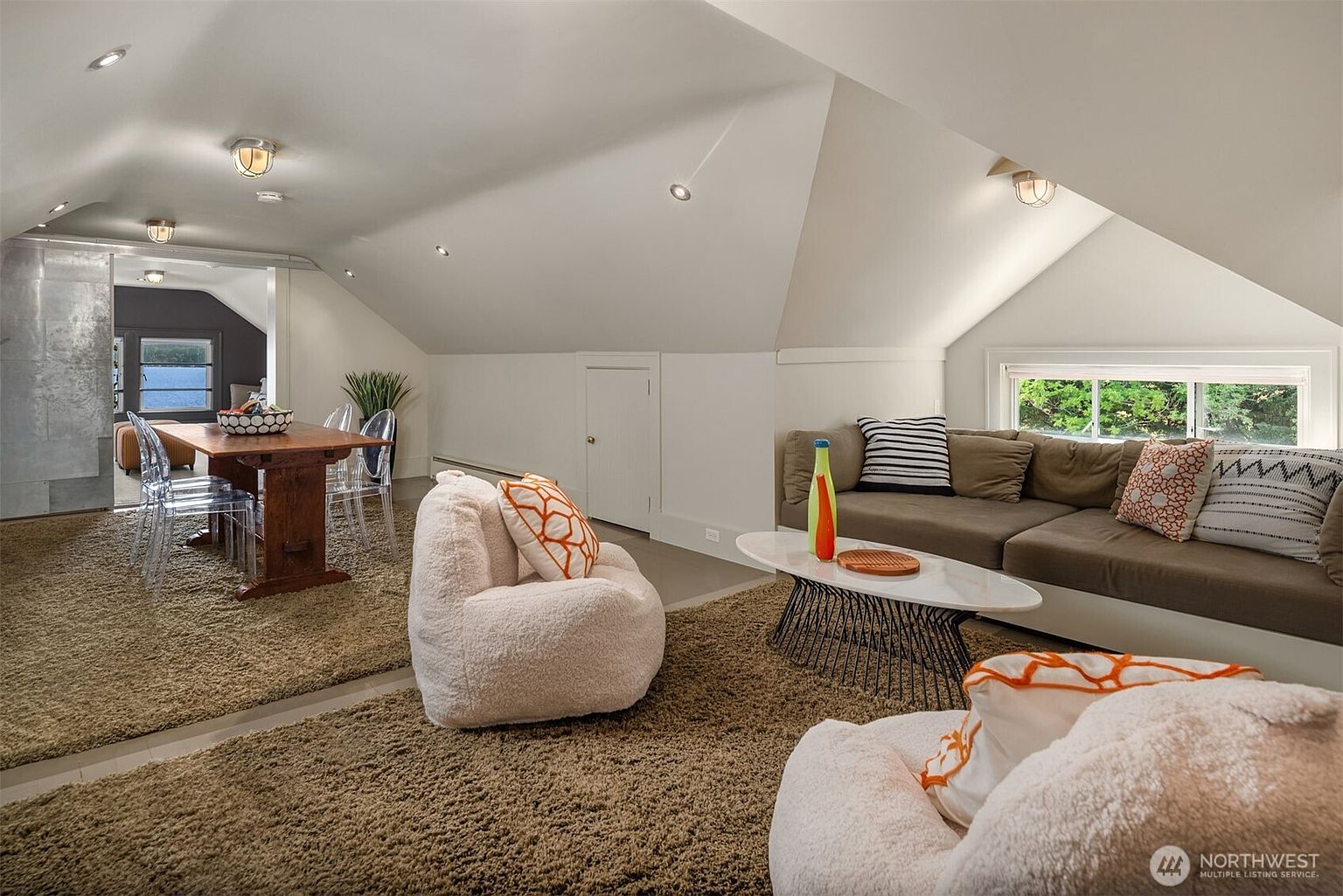 This spacious, open-concept attic living area features a cozy lounge space with a large sectional sofa and plush, textured armchairs set atop a thick, shaggy carpet. The room is illuminated by recessed lighting and natural light from a low-profile window, while a dining area with a rustic wooden table and transparent chairs is visible in the background. The slanted ceilings and neutral color palette create a warm, inviting atmosphere perfect for a versatile multi-use living space.