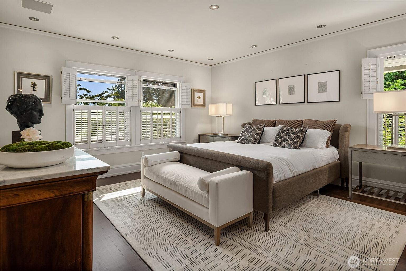 This elegant primary bedroom features a sophisticated neutral color palette, centered around a plush, upholstered bed frame and a matching white bench at the foot. The room is bathed in natural light from large windows adorned with classic plantation shutters, while a dark wood dresser with a decorative bust adds a touch of traditional charm. The space is finished with a textured area rug and minimalist wall art, creating a serene and luxurious atmosphere.