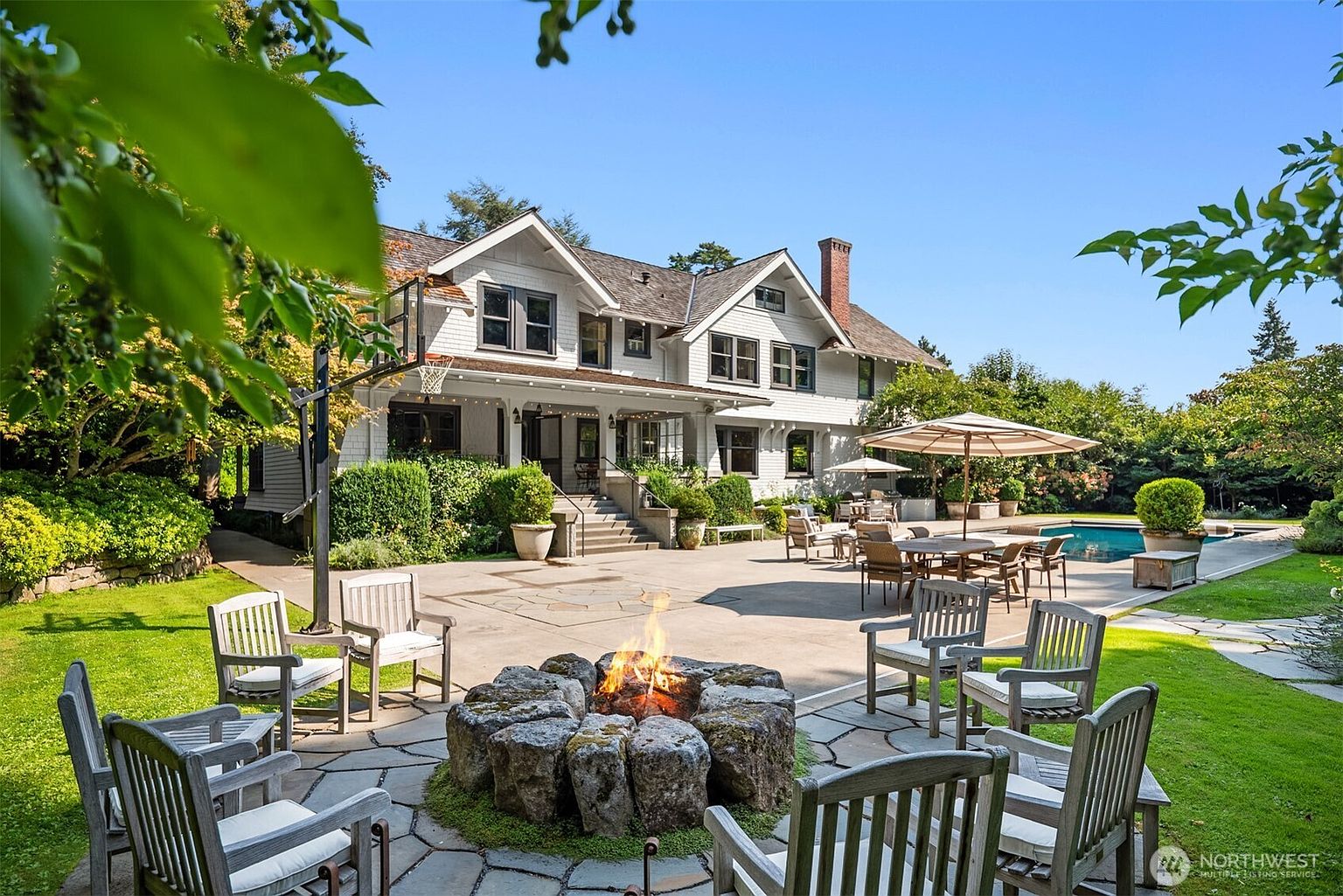 This expansive rear view of a traditional-style home showcases a beautifully landscaped backyard featuring a stone fire pit, a spacious patio, and a swimming pool. The scene is framed by lush greenery and mature trees, creating a serene and inviting outdoor living space perfect for entertaining. The perspective captures the seamless transition from the home's covered porch to the open-air recreational area.