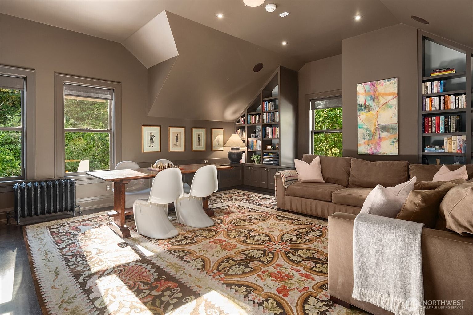 This cozy, multi-functional living room features a warm, earth-toned color palette with a large brown sectional sofa and a patterned area rug. The space includes a dedicated dining or game area with modern white chairs, built-in bookshelves, and framed artwork, all illuminated by natural light from multiple windows. The room's sloped ceiling and rich brown walls create an inviting, sophisticated atmosphere perfect for relaxation or study.
