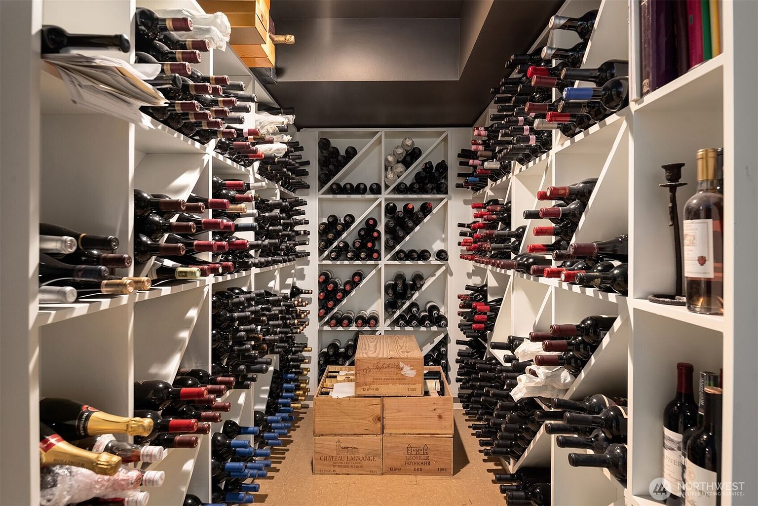 This sophisticated, climate-controlled wine cellar features extensive custom white shelving packed with a diverse collection of bottled wines. The space is organized with both individual bottle racks and wooden storage crates, including labels like Chateau Lagrange and Leoville Poyferre, creating an atmosphere of luxury and connoisseurship. The perspective is a centered, eye-level shot looking down the aisle, emphasizing the depth and abundance of the collection.