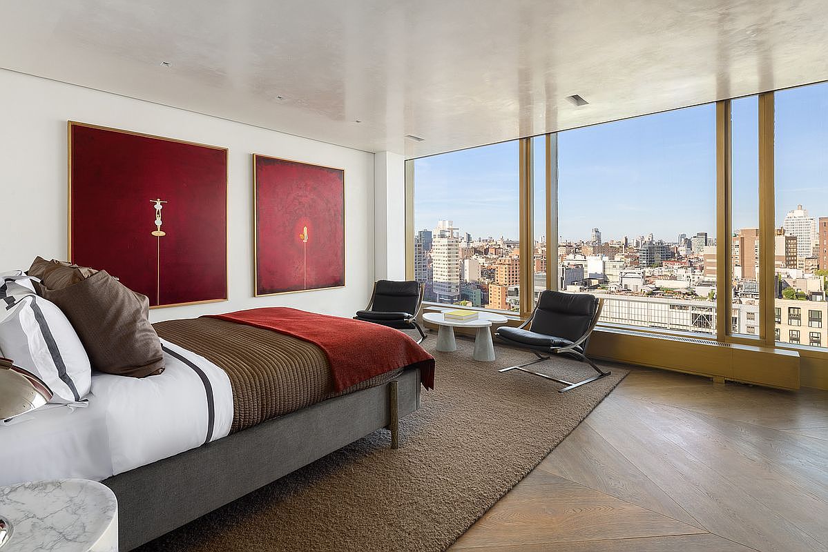 This sophisticated primary bedroom features a minimalist aesthetic with a neutral color palette accented by two large, bold red abstract art pieces above the bed. The room is defined by expansive floor-to-ceiling windows that provide a panoramic view of the city skyline, complemented by a pair of modern black leather lounge chairs and a small coffee table. The space is grounded by a textured area rug over warm-toned wood flooring, creating a luxurious and serene atmosphere.