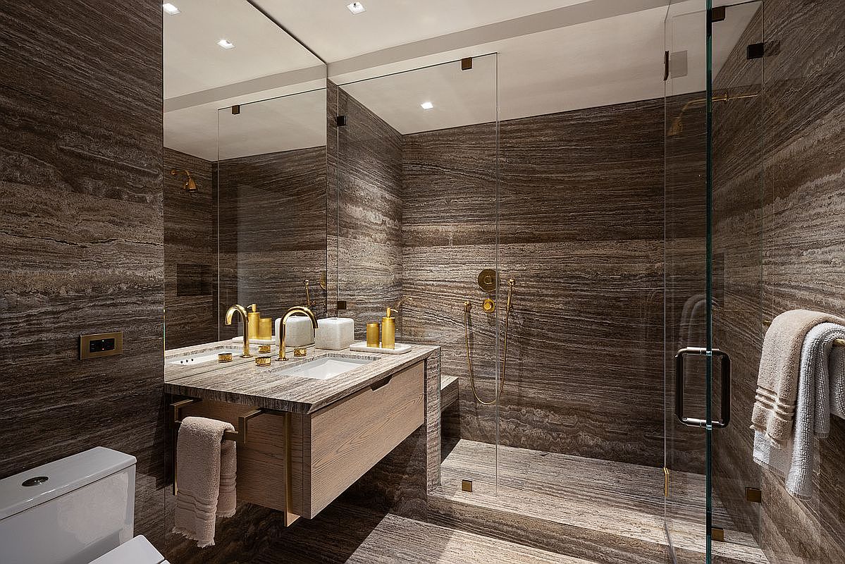 This luxurious bathroom features striking, horizontally-veined stone walls that create a cohesive and sophisticated aesthetic. The space includes a floating light-wood vanity with gold-toned fixtures, a walk-in shower enclosed by glass panels, and a modern toilet. The perspective captures the entire vanity area and shower entrance, highlighting the high-end finishes and seamless design.