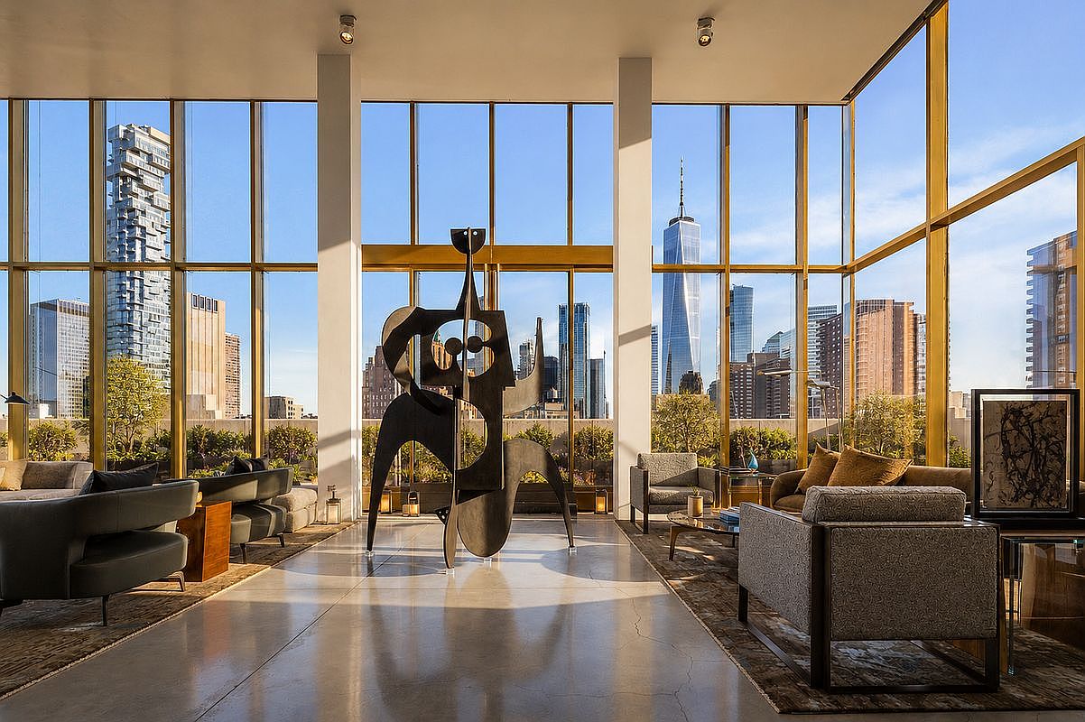 This sophisticated, high-rise living space features expansive floor-to-ceiling windows that offer a panoramic view of the New York City skyline, including the One World Trade Center. The room is anchored by a large, abstract metal sculpture in the center, surrounded by contemporary seating arrangements on plush area rugs. The polished concrete floors and minimalist architectural columns create a sleek, gallery-like atmosphere that emphasizes the dramatic urban backdrop.
