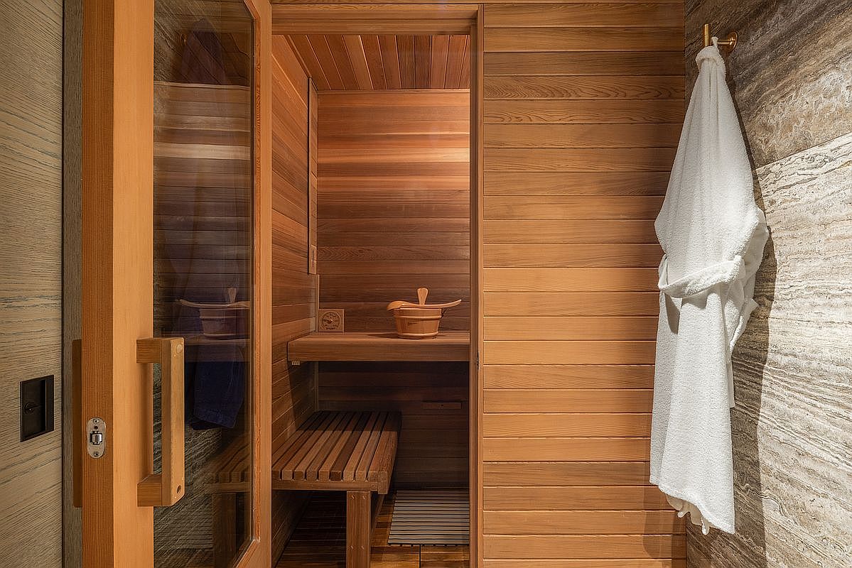 This image showcases a custom-built sauna featuring warm-toned wood paneling throughout the interior and exterior walls. A glass-paneled door provides a clear view into the sauna, which is equipped with a wooden bench, a small bucket, and a ladle, creating a serene spa-like atmosphere. To the right, a plush white bathrobe hangs on a hook against a textured stone wall, adding a touch of luxury to the space.