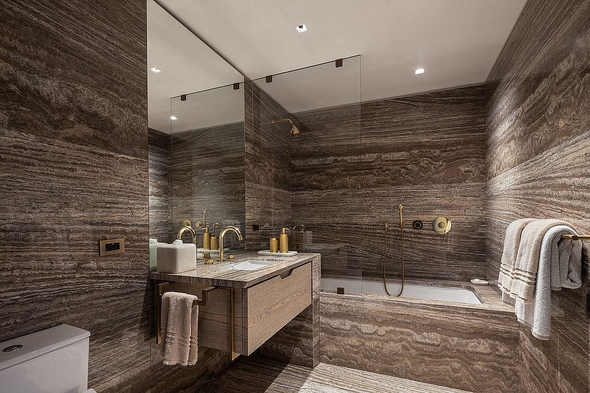 This luxurious bathroom is defined by its dramatic, horizontal-veined stone walls that create a cohesive and sophisticated aesthetic. The space features a floating wood vanity with a stone countertop, a glass-enclosed shower and tub combination, and elegant gold-toned fixtures. A large mirror reflects the space, enhancing the sense of depth and highlighting the high-end, modern design.