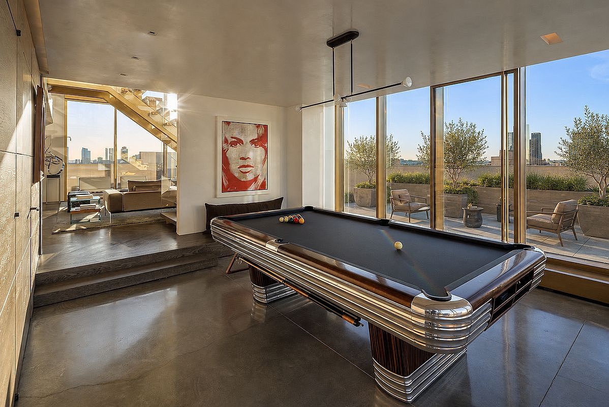 This sophisticated game room features a prominent, high-end pool table with a dark felt surface as the central focal point. The space is defined by polished concrete floors, a striking piece of pop art on the wall, and expansive floor-to-ceiling windows that provide a seamless transition to an outdoor terrace. A raised, carpeted seating area is visible in the background, creating a multi-level layout that enhances the open, airy atmosphere of this luxury residence.