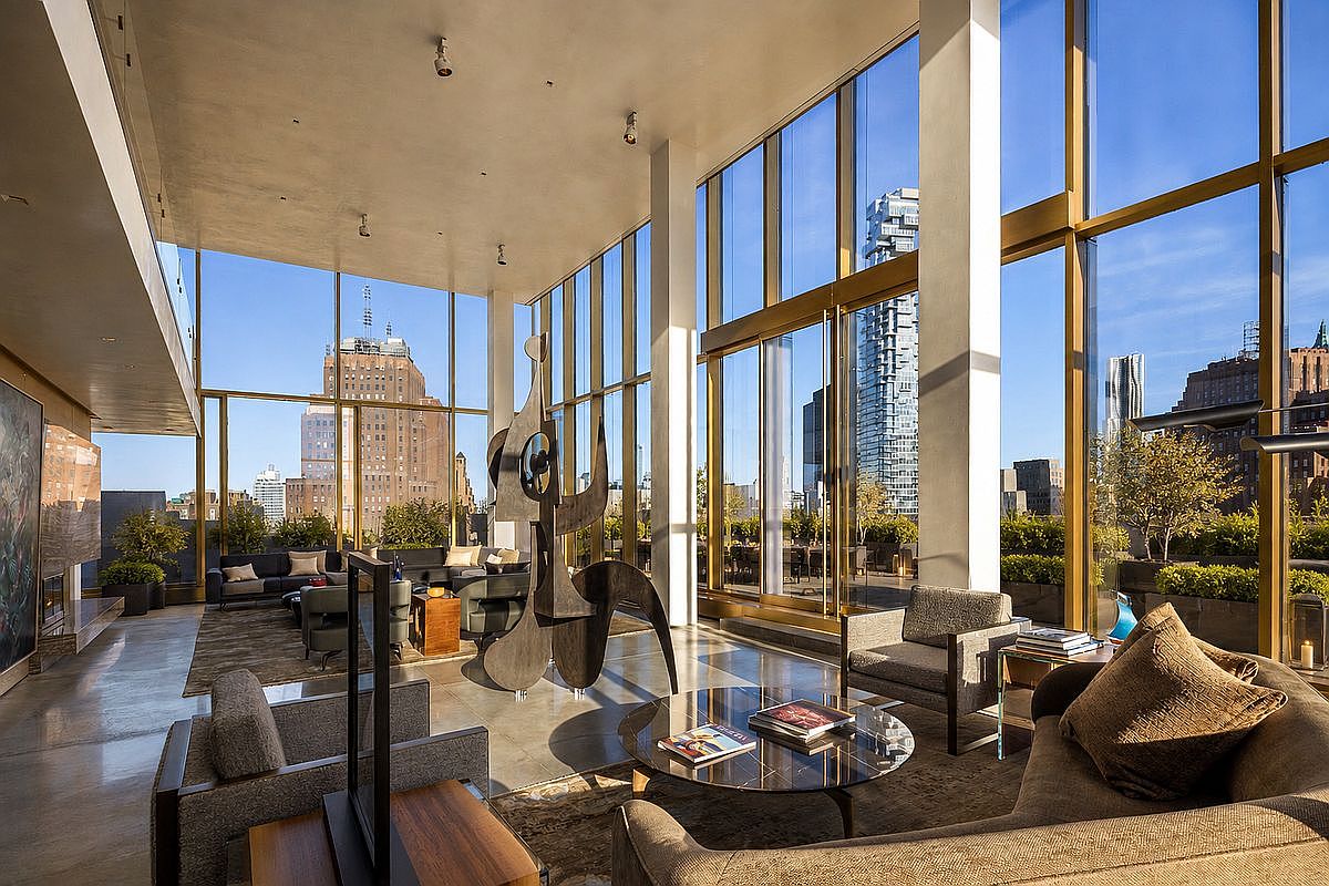 This expansive, high-ceilinged living room features floor-to-ceiling windows that offer panoramic city views and flood the space with natural light. The interior is anchored by a large, abstract metal sculpture and furnished with contemporary seating arrangements, including plush sofas and armchairs arranged around a circular coffee table. The polished concrete flooring and minimalist aesthetic create a sophisticated, modern atmosphere suitable for luxury urban living.