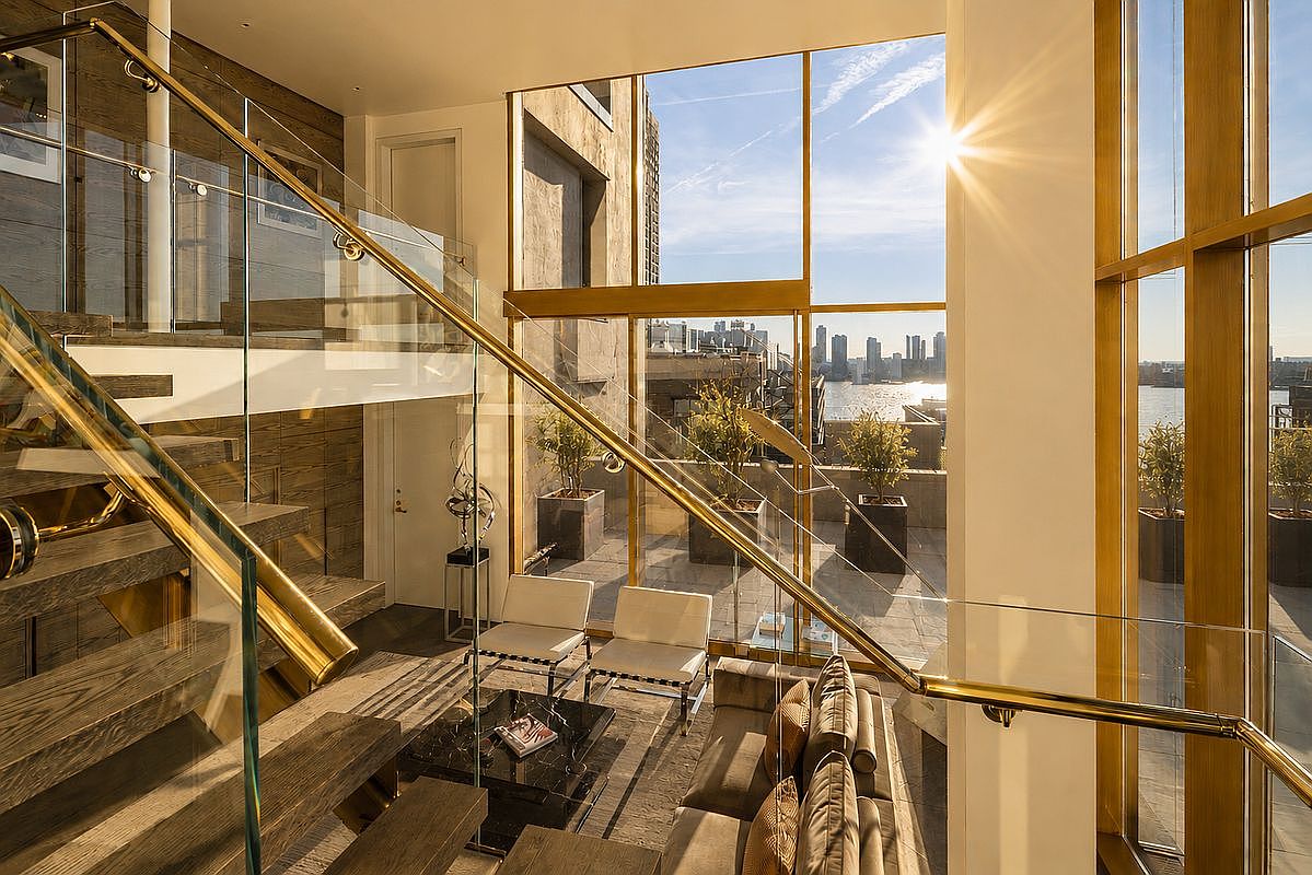 This sophisticated, multi-level living space features a dramatic staircase with glass railings and wood treads that overlooks a seating area below. The room is defined by expansive, floor-to-ceiling windows that frame a panoramic city and waterfront view, bathing the interior in warm, natural sunlight. The decor includes modern white armchairs, a dark coffee table, and a plush sofa, creating an elegant and airy atmosphere.