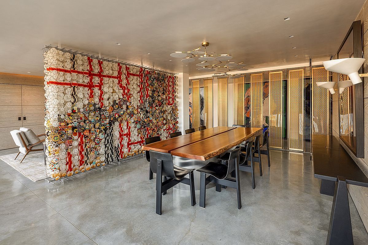 This sophisticated dining area features a long, live-edge wood table centered in the room, surrounded by modern black chairs. A striking, textured art installation serves as a room divider, while a series of metallic, grid-patterned screens line the back wall. The space is illuminated by a contemporary multi-tiered chandelier and sleek wall-mounted lighting, creating a refined and artistic atmosphere.