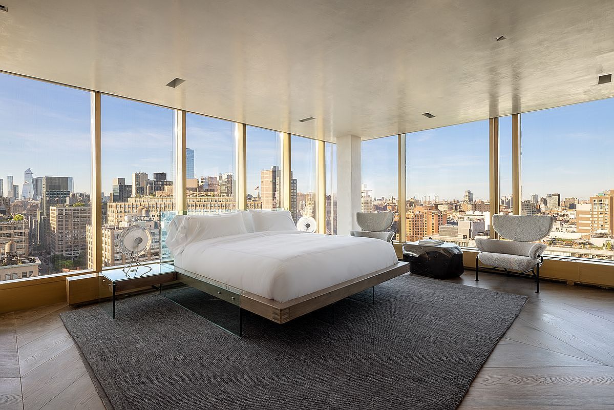 This expansive primary bedroom features a minimalist aesthetic centered around a platform bed positioned to take full advantage of the panoramic city views. The room is enclosed by floor-to-ceiling windows that wrap around the space, showcasing a high-rise urban skyline. A dark, textured area rug anchors the bed, while two modern armchairs provide a comfortable seating area, all set against a backdrop of light wood flooring and a smooth, neutral ceiling.